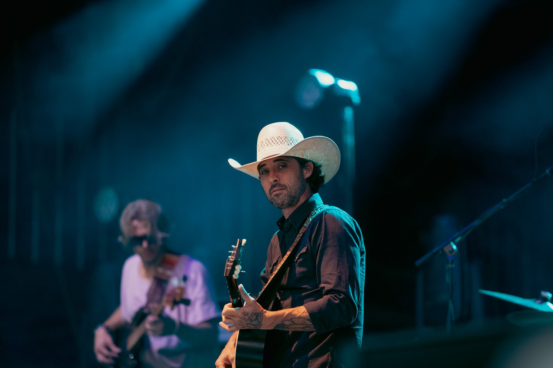 Musicians performing on stage, one wearing a cowboy hat and playing an acoustic guitar, with another blurred musician in the background.