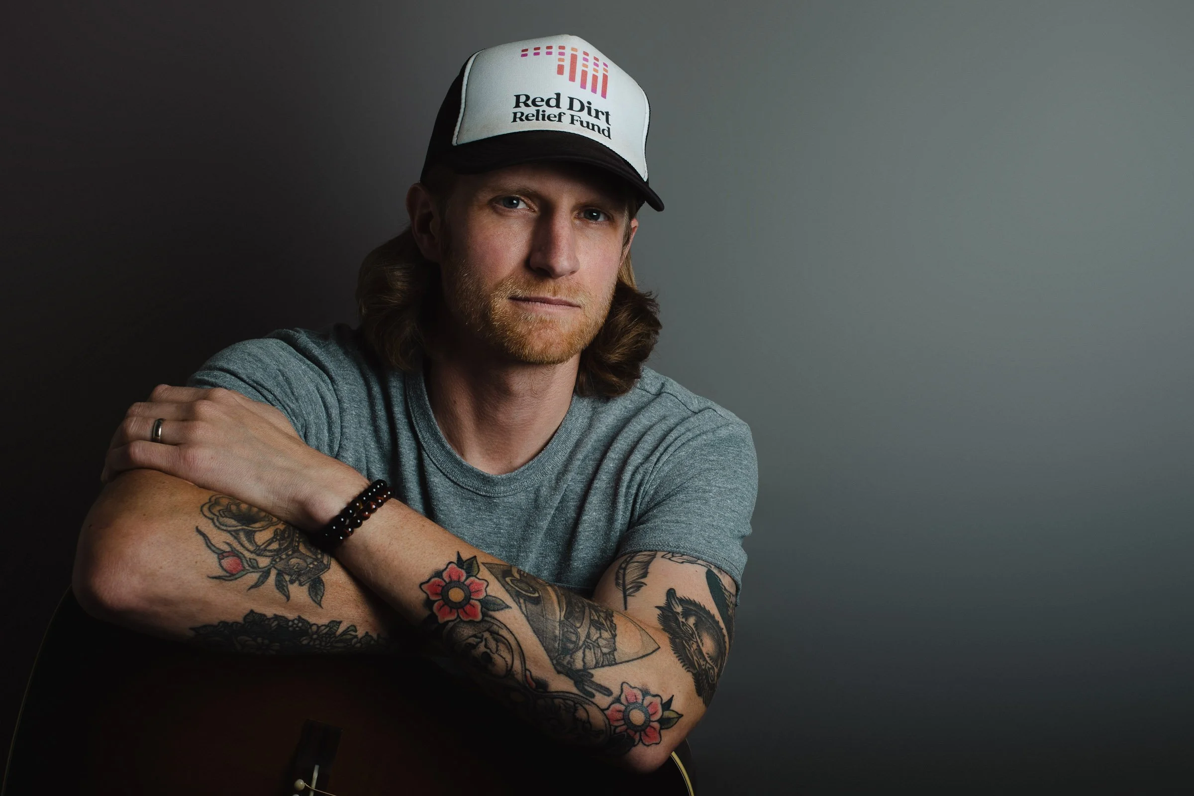 A man with long hair and a beard wearing a gray t-shirt and a trucker hat with the logo 'Red Dirt Relief Fund' poses with his arms crossed, revealing tattoos on his left arm, against a plain background.