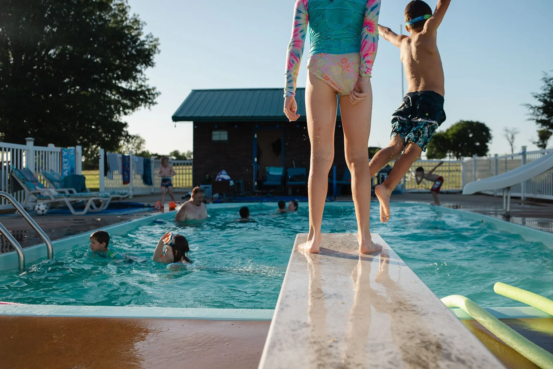Children and adults swimming and playing at an outdoor pool on a sunny day, with a small building and trees in the background.