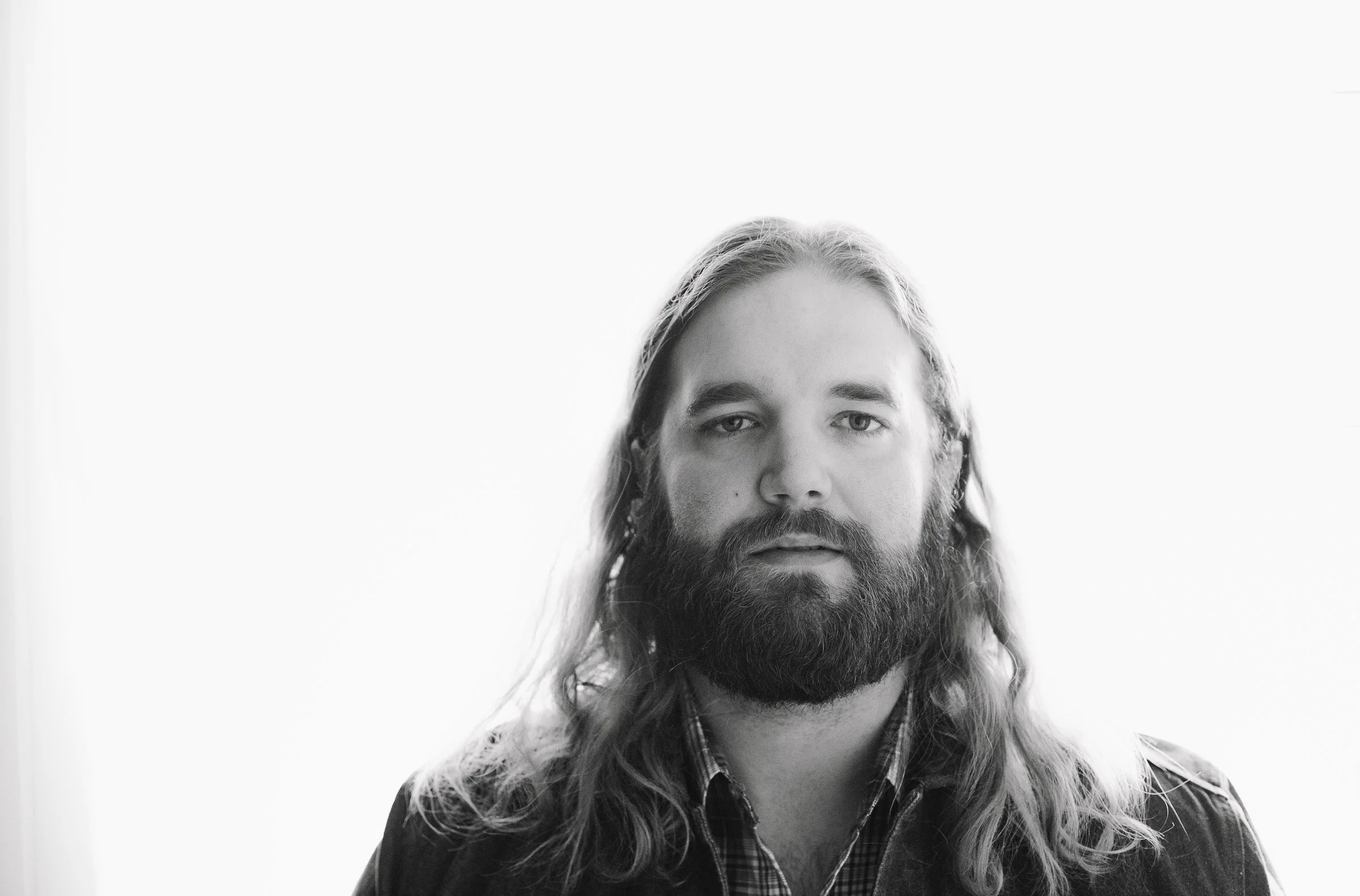 Black and white portrait of a man with long hair and a beard, wearing a plaid shirt under a jacket, looking directly at the camera with a neutral expression.