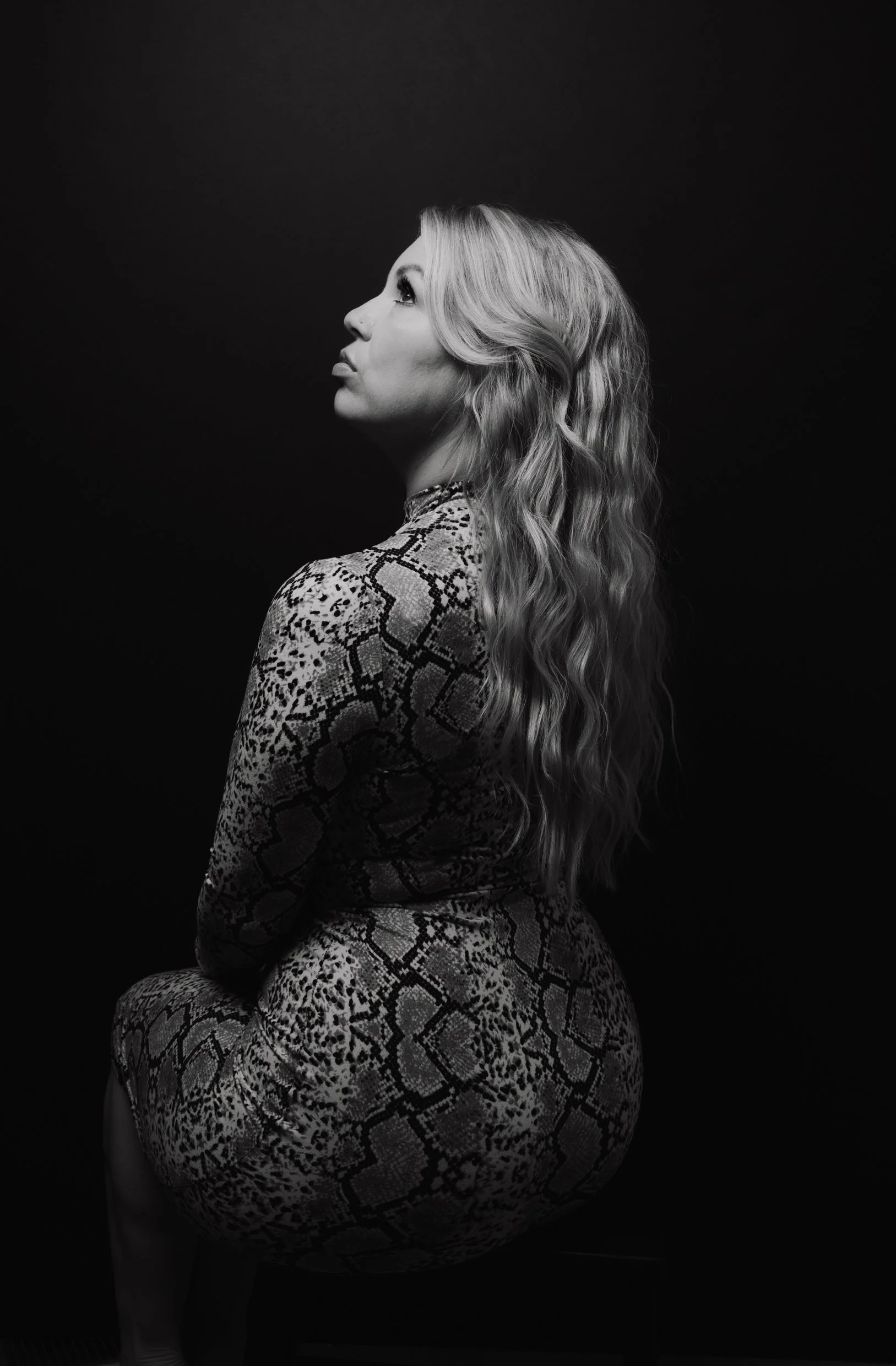 Black and white portrait of a woman with long wavy hair, wearing a snakeskin-patterned dress, sitting sideways with eyes looking up.