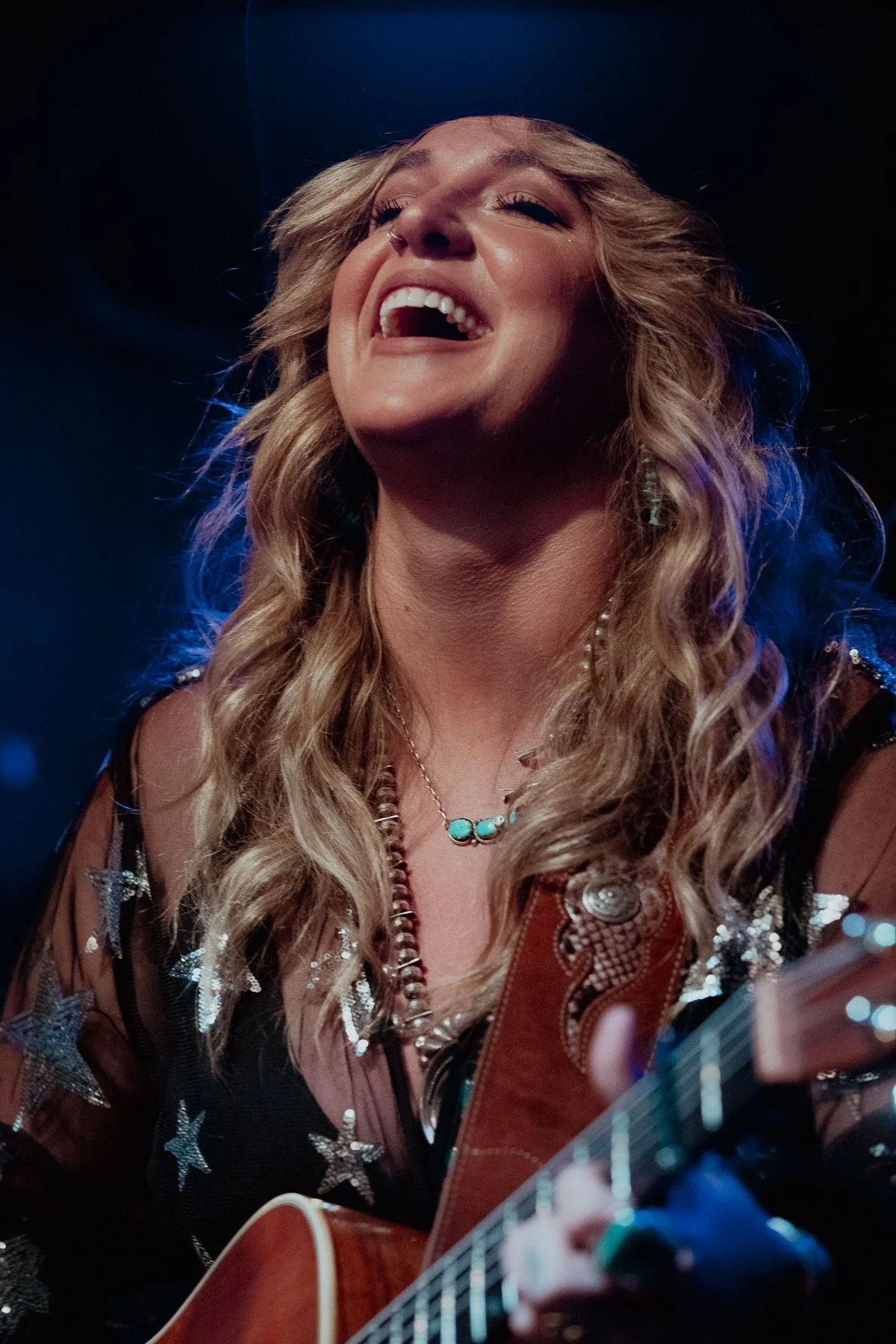 A woman with long blonde wavy hair is smiling and singing while playing an acoustic guitar. She is wearing a black top with star patterns, a pearl and turquoise necklace, and has earrings.