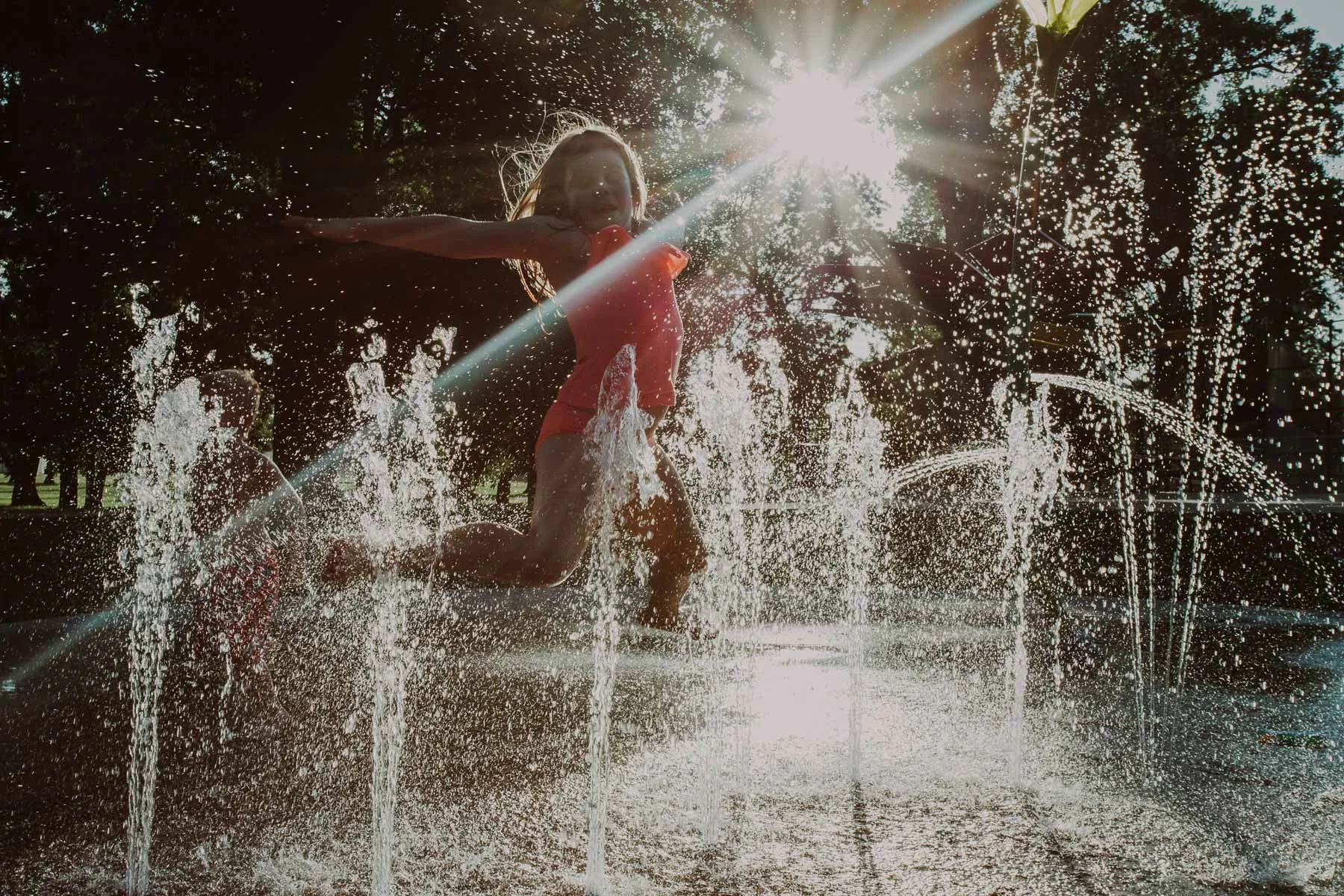 Children playing and running through a splash pad or water fountain in a park, with the sun shining brightly in the background.