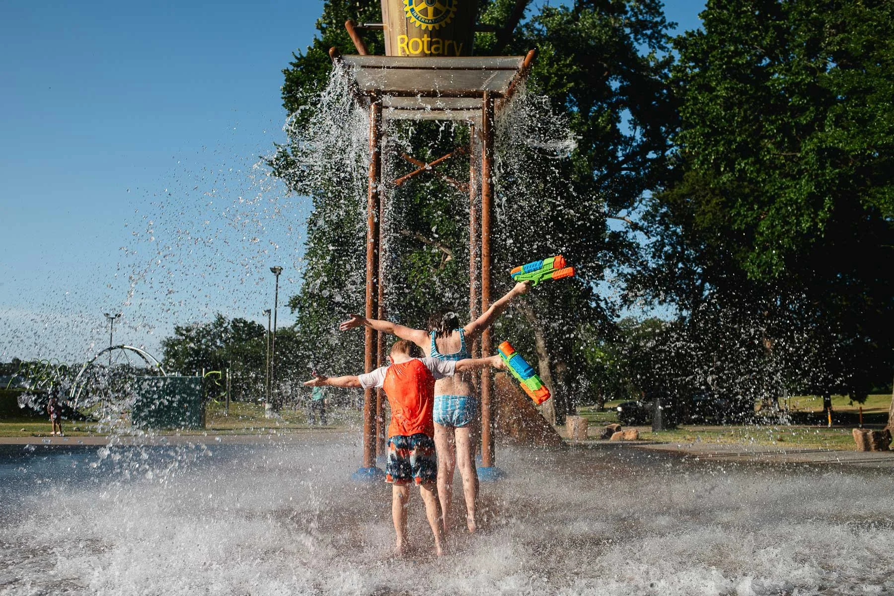 Children playing in water spray at a splash pad with water guns, under a wooden structure with a Rotary International sign, surrounded by green trees on a sunny day in a park.