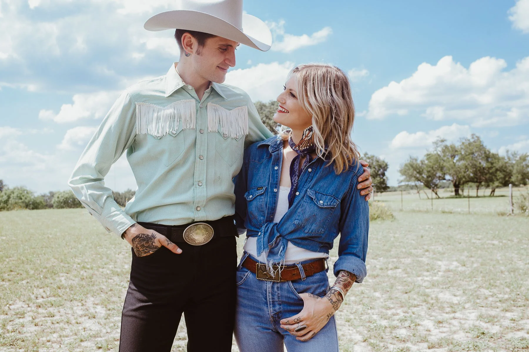 A man and woman dressed in Western attire standing outdoors on a grassy field with trees and blue sky in the background. They are looking at each other and smiling, with the man wearing a cowboy hat and the woman with tattoos visible on her forearms 