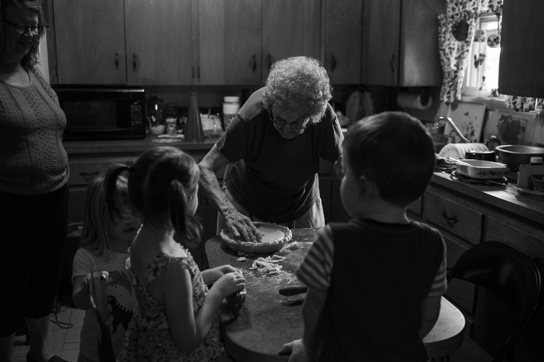 An elderly woman rolling out pie dough at a kitchen table, surrounded by three young children and a woman. The kitchen has wooden cabinets and various dishes on the counter.