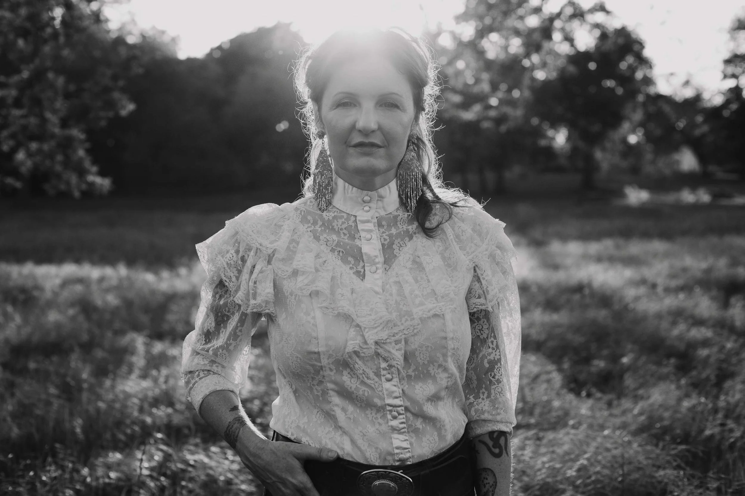 A woman standing outdoors in a field with trees in the background, backlit by the sun, wearing a lace blouse, earrings, and belt, with tattoos visible on her arm.