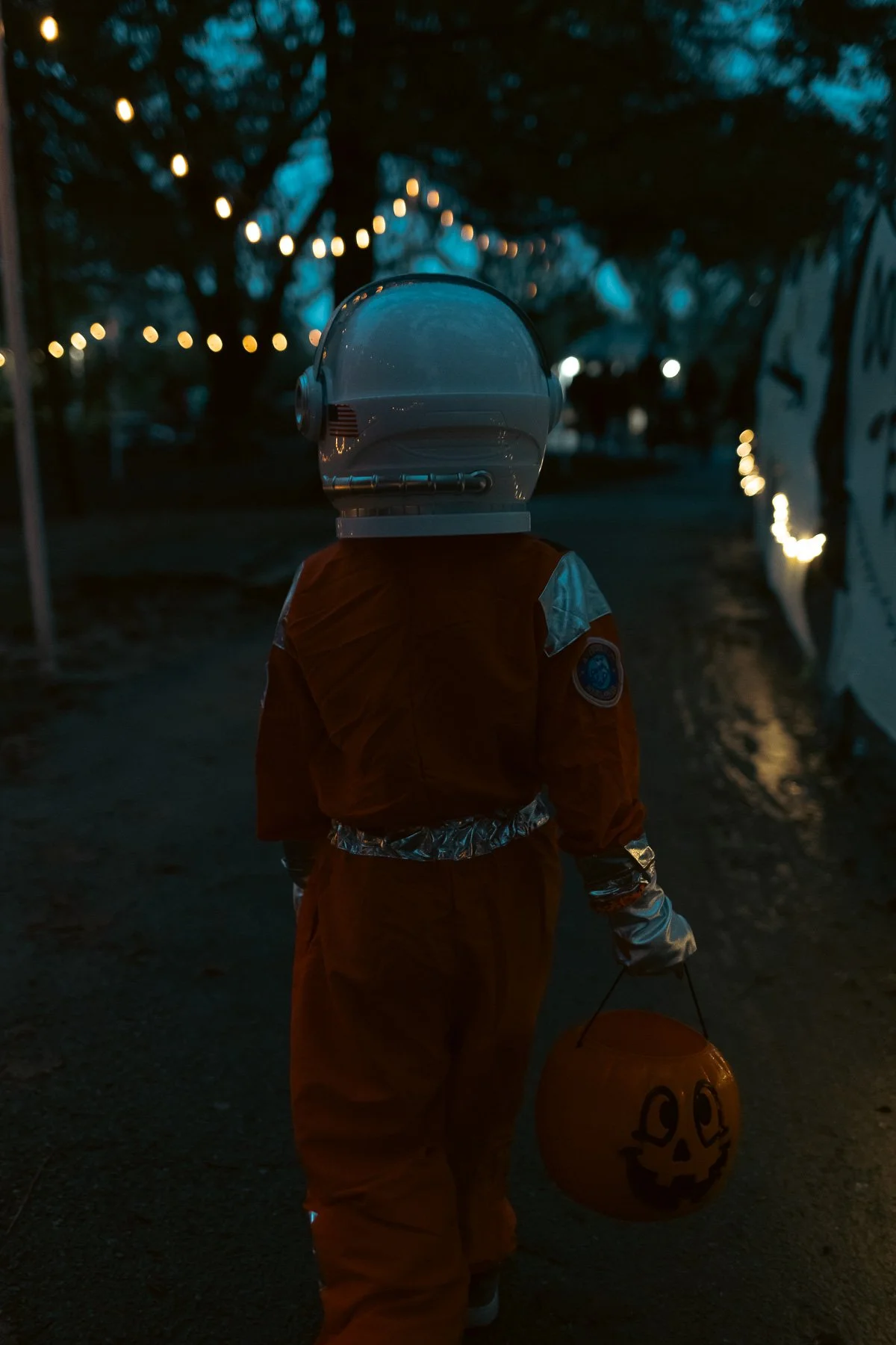 A child dressed in an orange spacesuit holding a pumpkin-shaped Halloween bucket walking outdoors at dusk, with string lights and trees in the background.