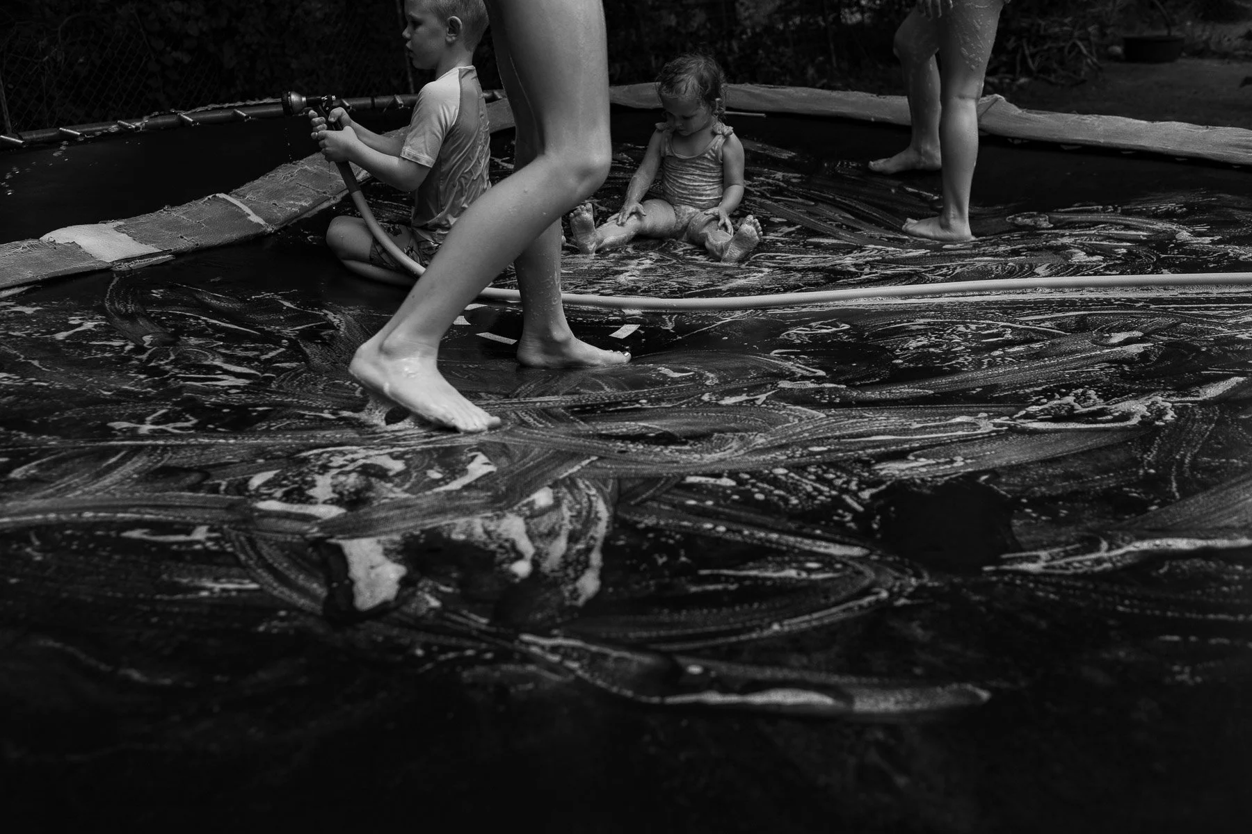 Children playing in a soapy, bubble-filled backyard pool with some standing, sitting, and holding a hose.
