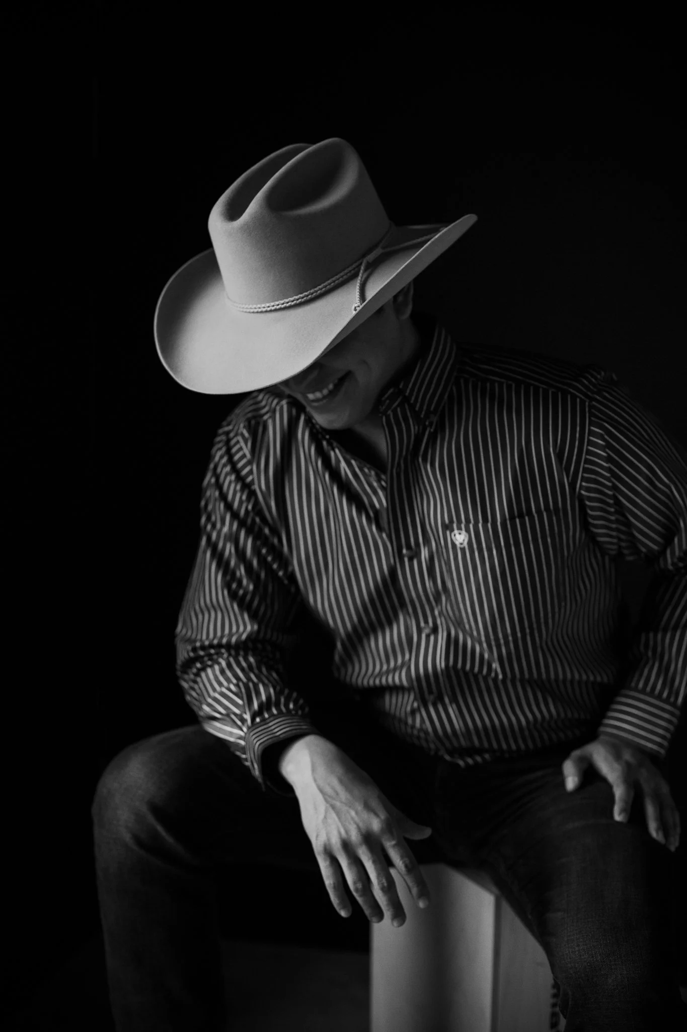 A person wearing a striped shirt and a wide-brimmed hat, sitting and smiling with their head tilted down, in black and white.