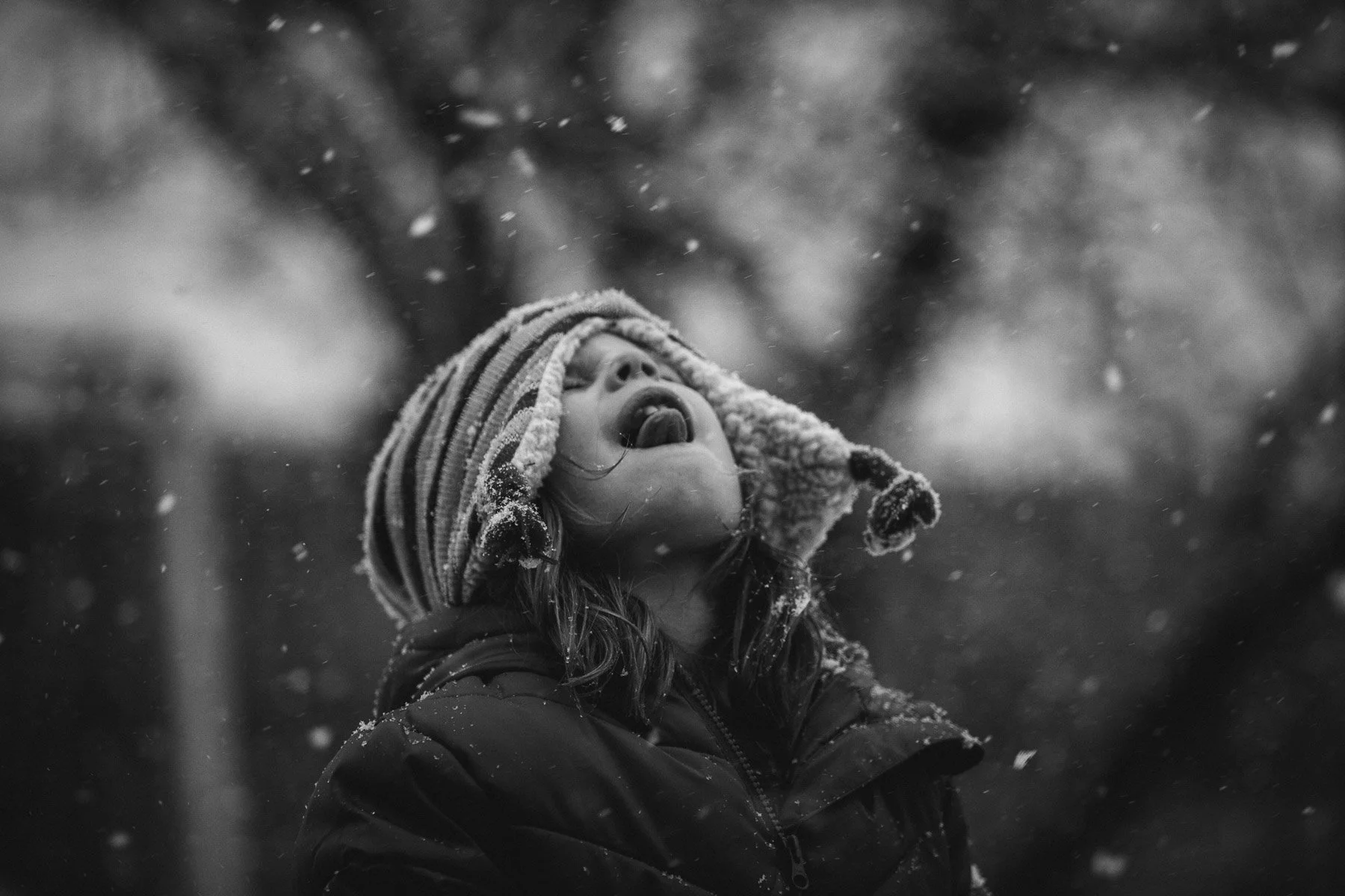 A girl in winter clothing, wearing a striped knit hat with ear flaps, stands outdoors in snow, looking up with her mouth open and tongue out, surrounded by falling snow.