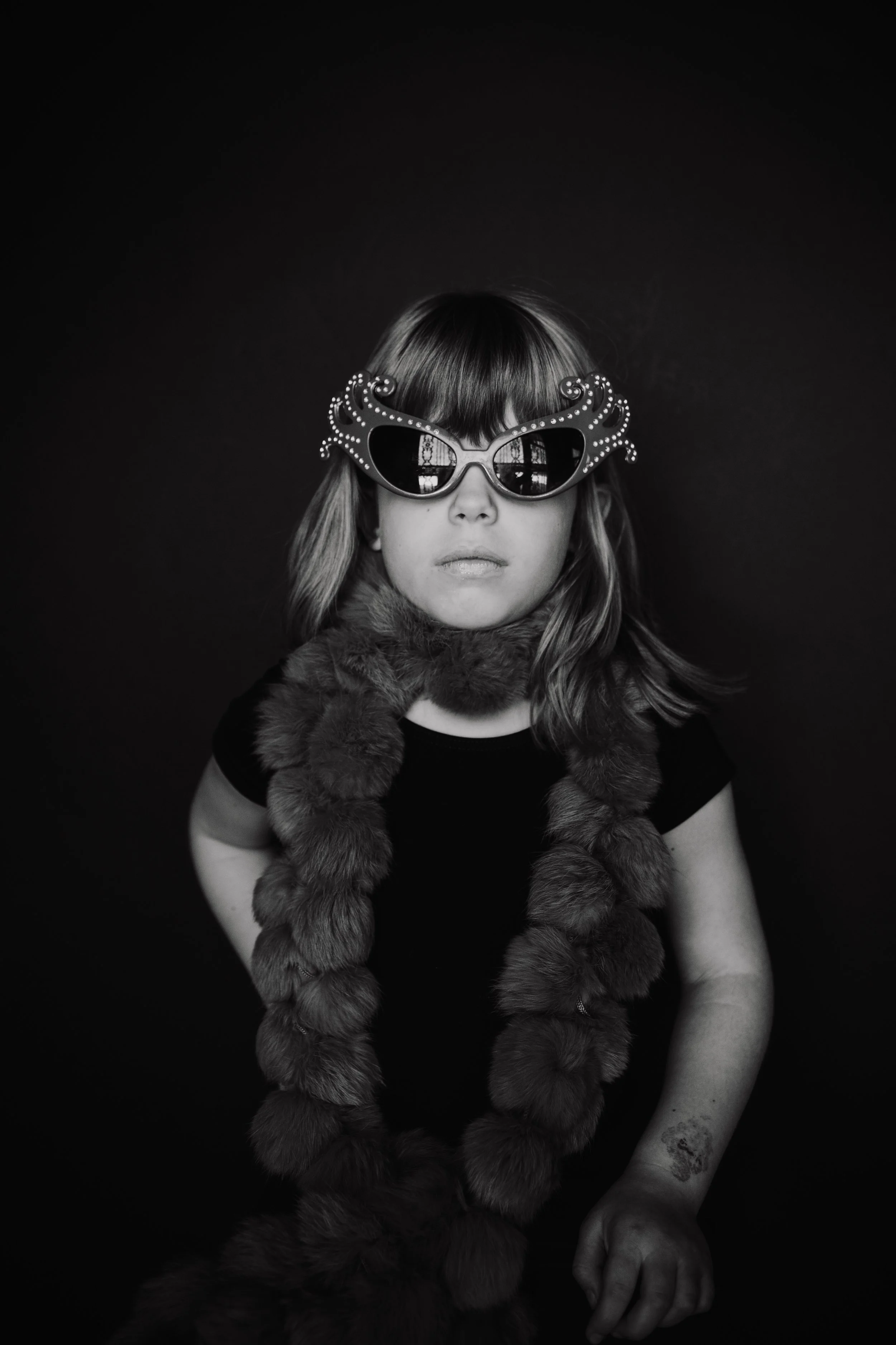 Black and white photo of a girl wearing large sunglasses with decorated frames, a fur scarf, and a black shirt. She has straight hair with bangs and is posed against a plain dark background.