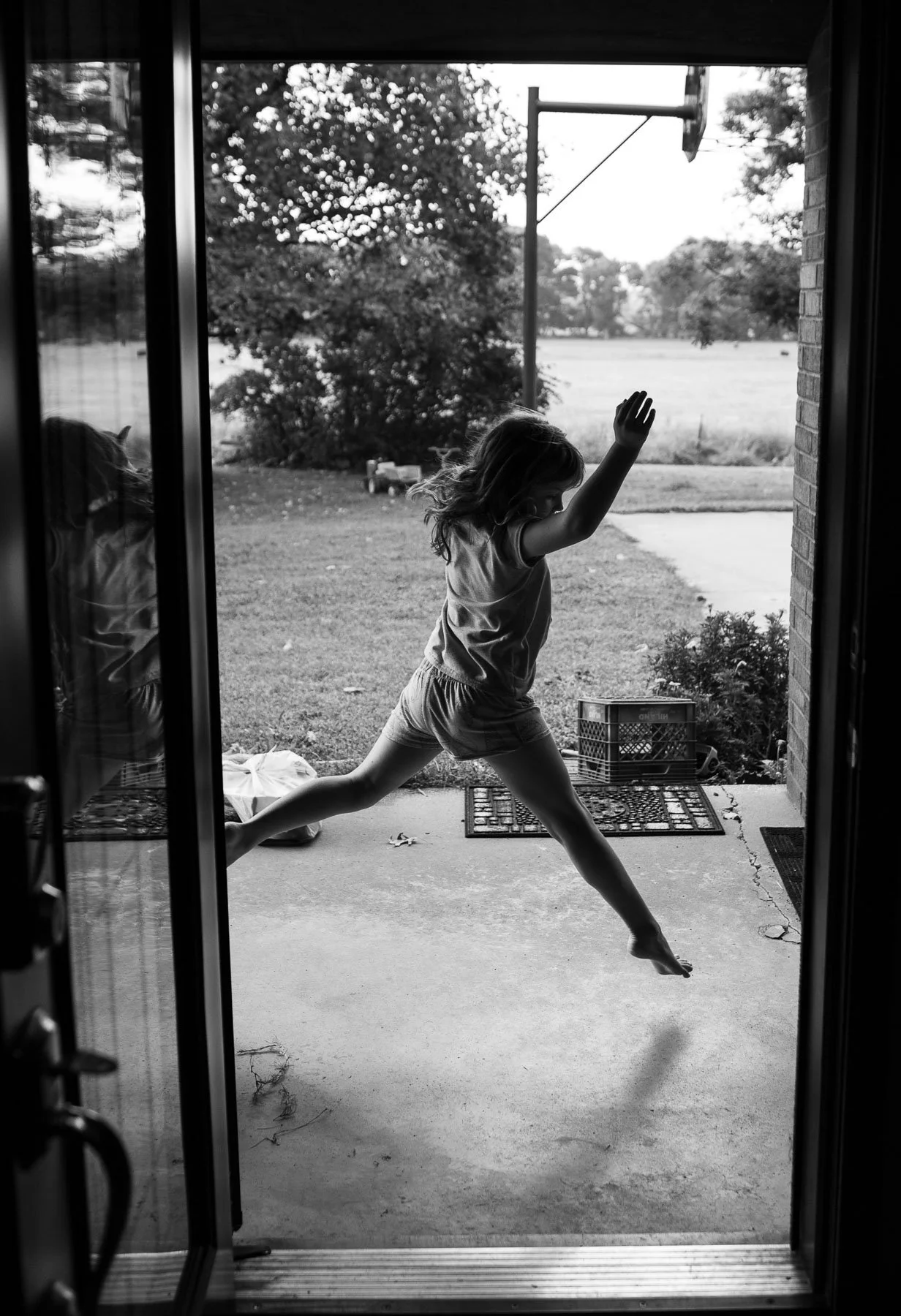 A young girl is jumping and reaching out with one arm while standing outside a glass door. The photo is taken from indoors, overlooking a backyard with trees, a basketball hoop, and a grassy field.