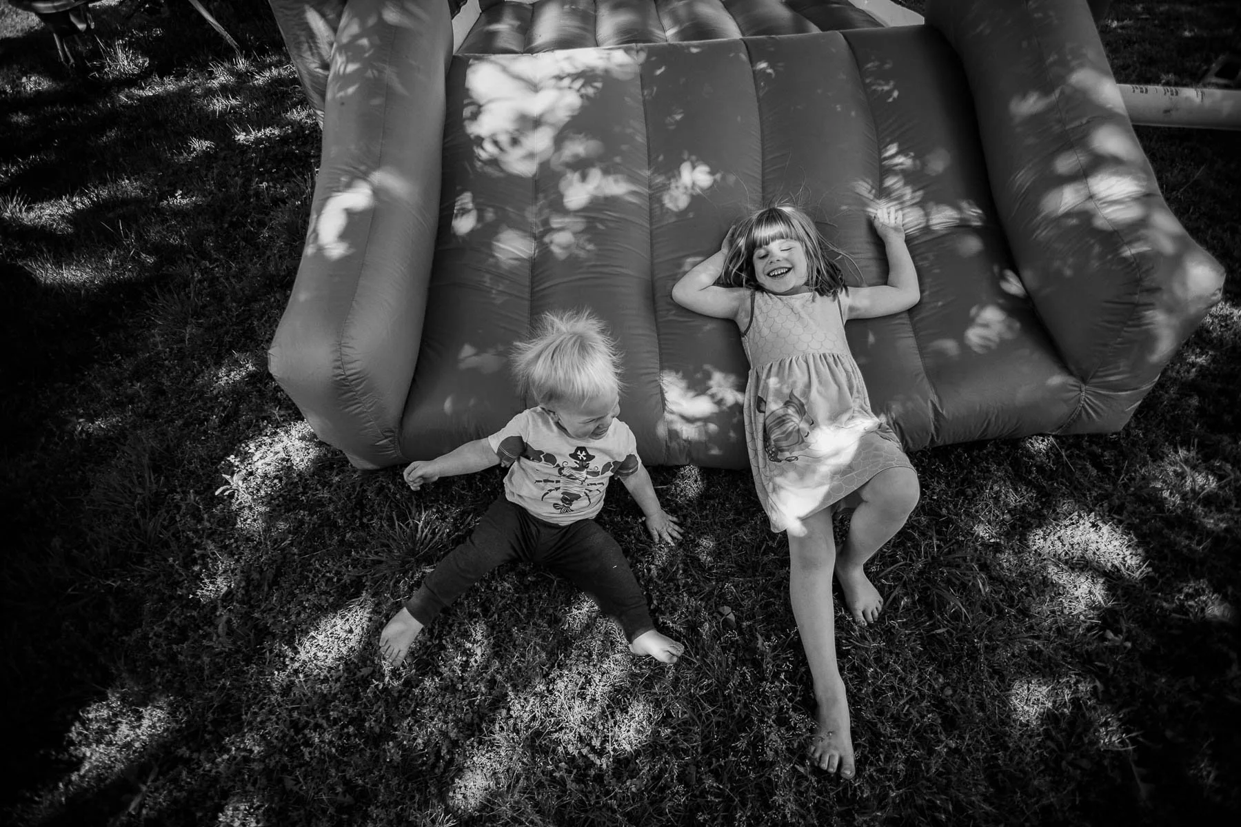 Two children, a girl and a younger boy, are lying on grass next to an inflatable sofa. The girl is smiling with her eyes closed, her arms raised, and is wearing a dress. The boy is looking down, with one arm touching the ground and the other slightly