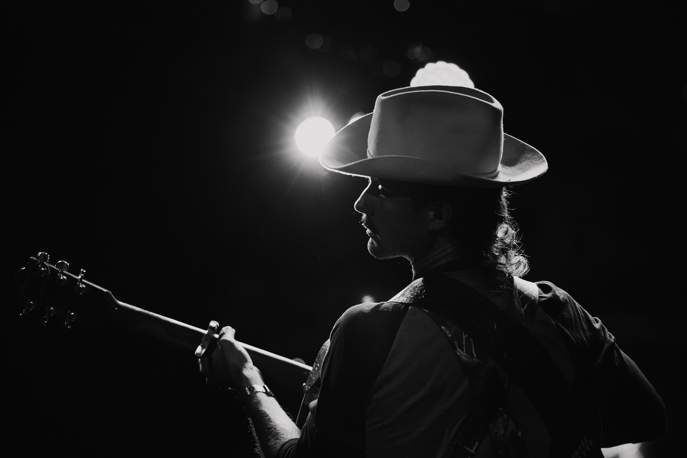 Black and white silhouette of a person wearing a cowboy hat and sunglasses, playing a guitar against a dark background with a bright light source behind them.
