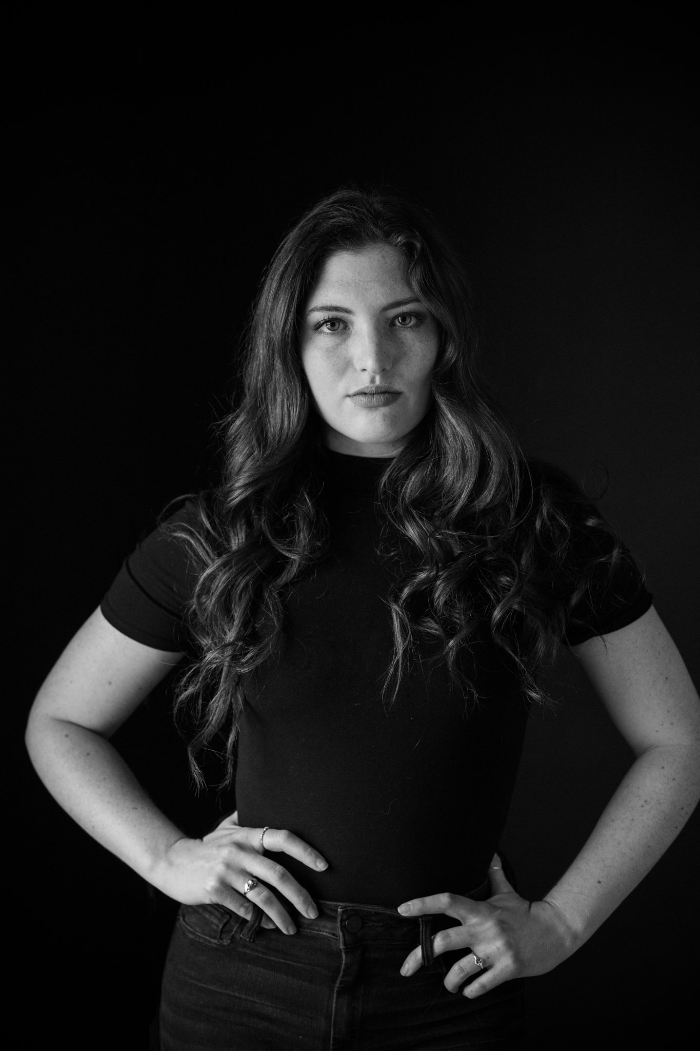 Black and white portrait of a woman with long wavy hair, wearing a fitted black t-shirt, standing against a dark background with her hands on her hips.