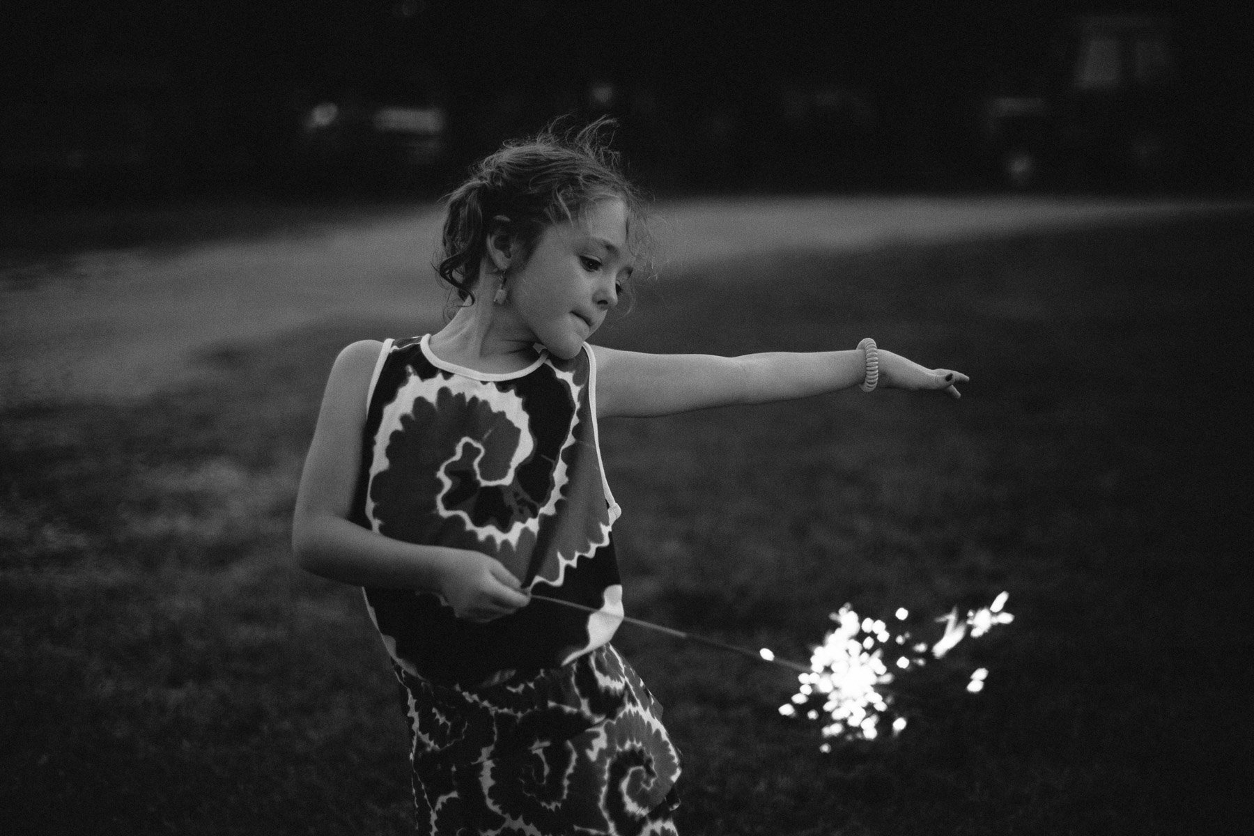 A young girl in a patterned dress plays with a sparkler in a dark, outdoor setting.