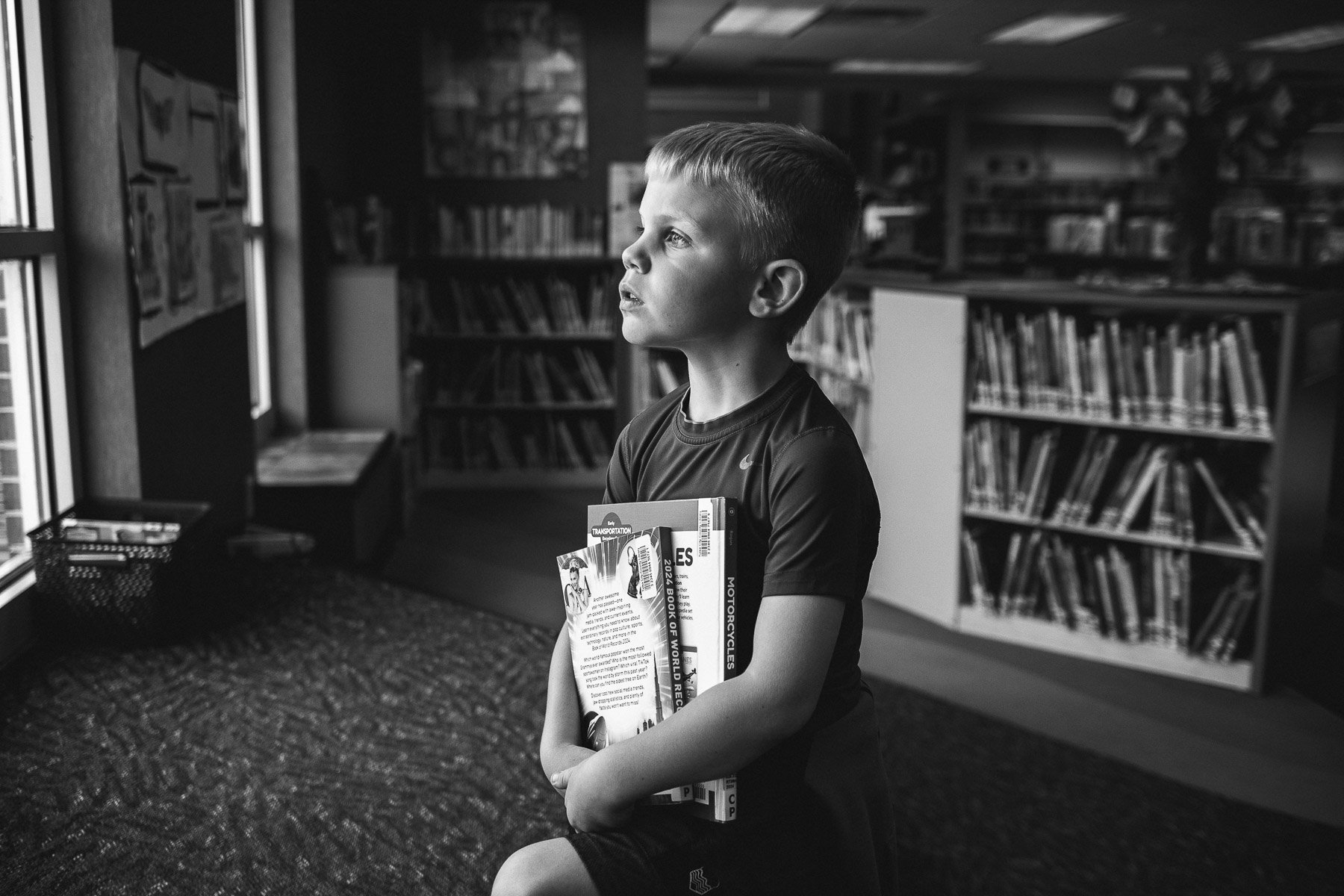 A young boy standing in a library holding books against his chest. He is looking to his left with a thoughtful expression. Bookshelves filled with books are visible in the background, and sunlight is coming through a window on the left.