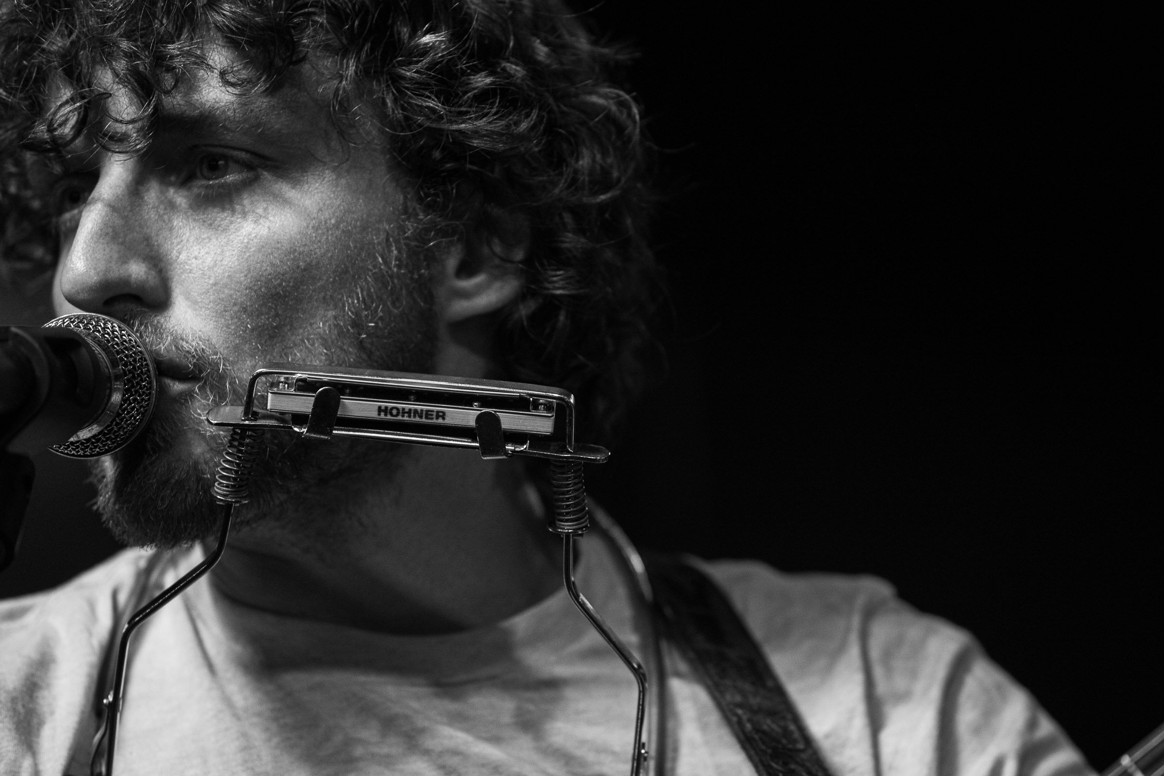 Black and white photo of a man with curly hair and a beard singing into a microphone while playing a harmonica.