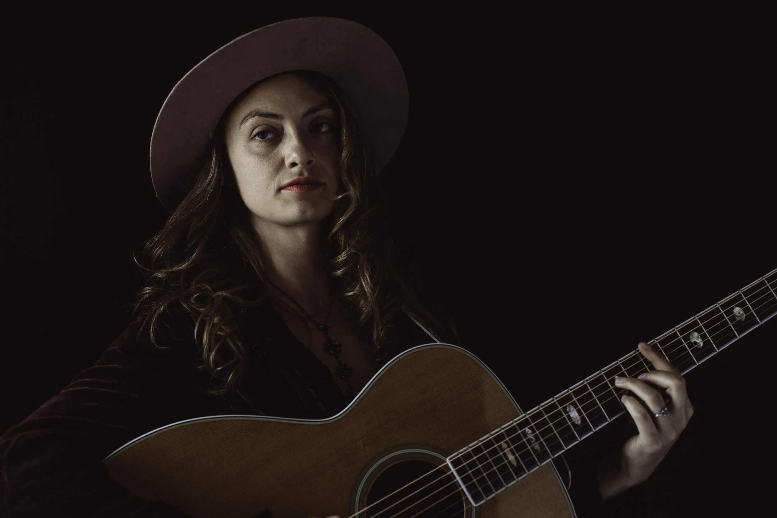 A woman in a hat holding an acoustic guitar against a dark background.