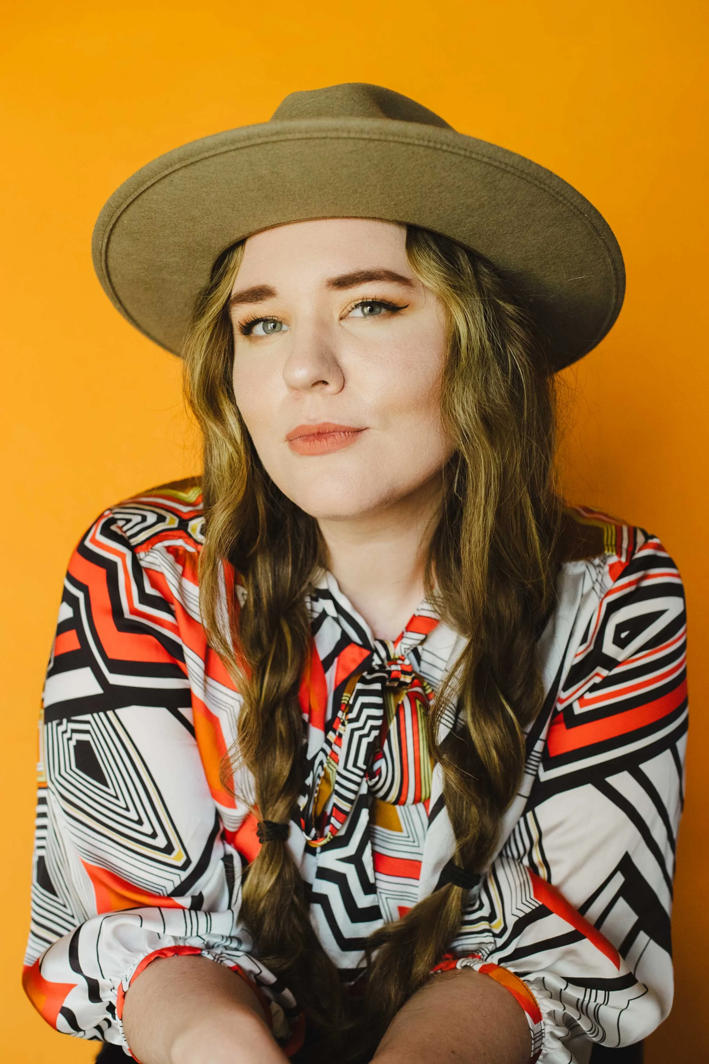 A woman wearing a wide-brimmed tan hat, a geometric patterned blouse with red, black, white, and yellow colors, and sporting long wavy hair with braids, against an orange background.
