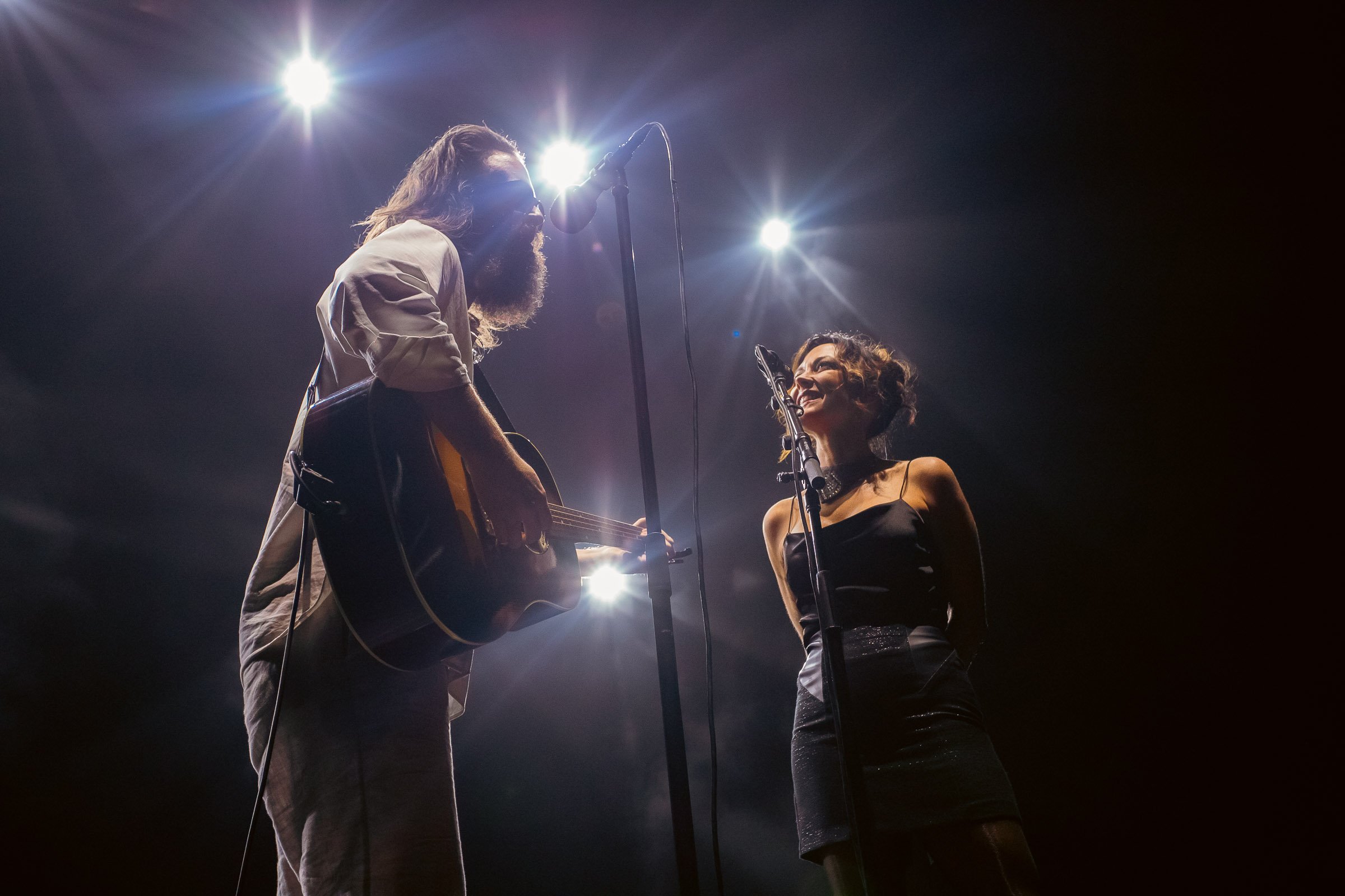 A male musician with long hair and a beard playing an acoustic guitar at a concert, and a female singer with curly hair wearing a black dress singing into a microphone, illuminated by stage lights.