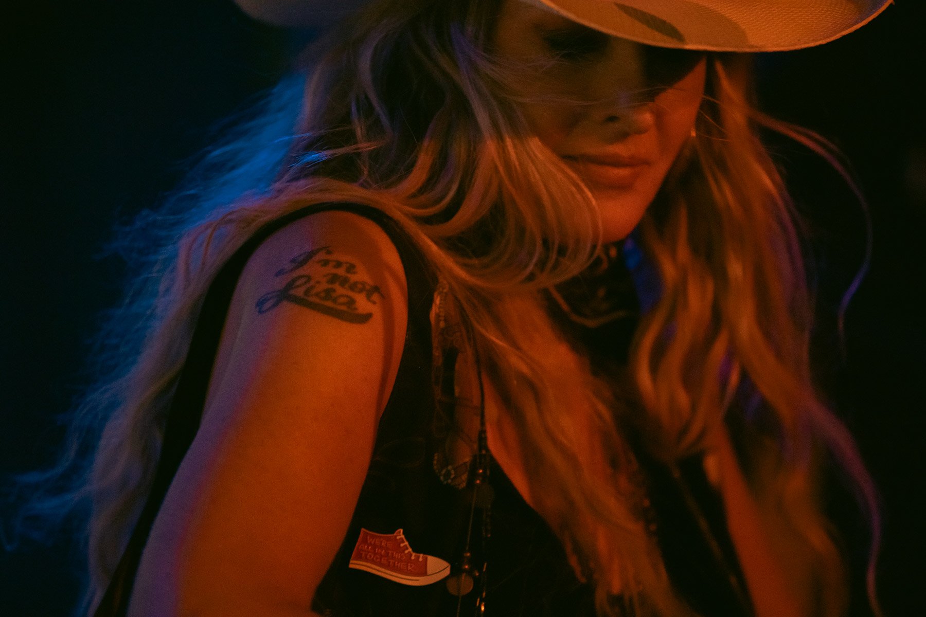 A woman with long, wavy blonde hair, wearing a western hat and a sleeveless shirt, slightly smiling, with colorful lighting on her face; showing a tattoo on her upper arm that reads "To the best" and other writings.