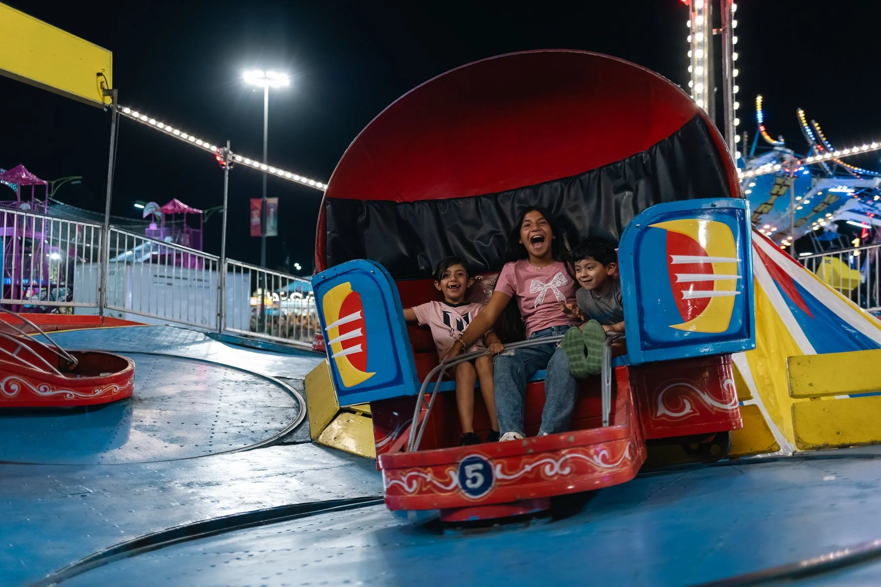 A family of three kids riding a colorful carnival ride at night, all showing excitement and joy.