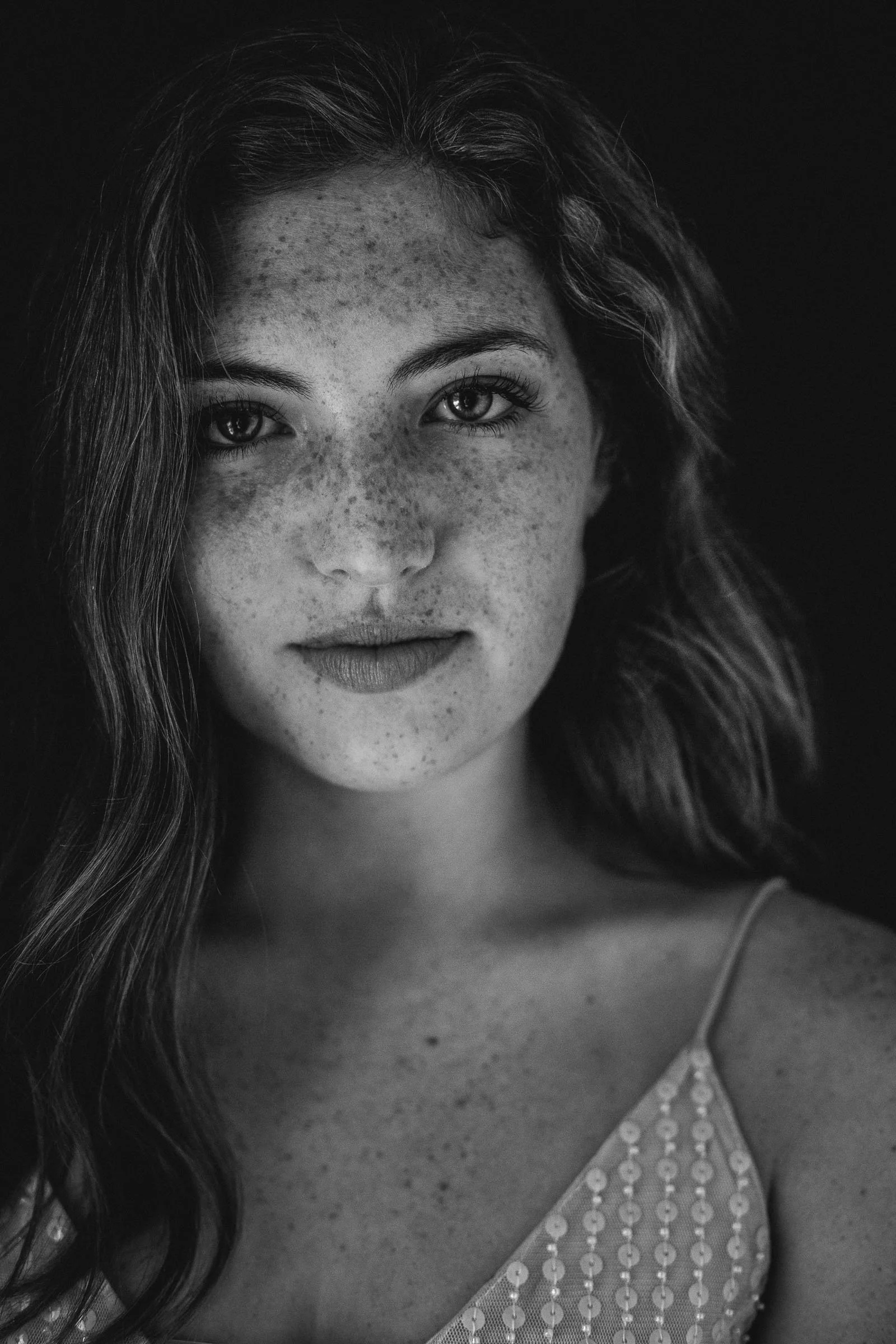 Close-up black and white portrait of a woman with long, wavy hair and freckles, wearing a sleeveless top with sequins.