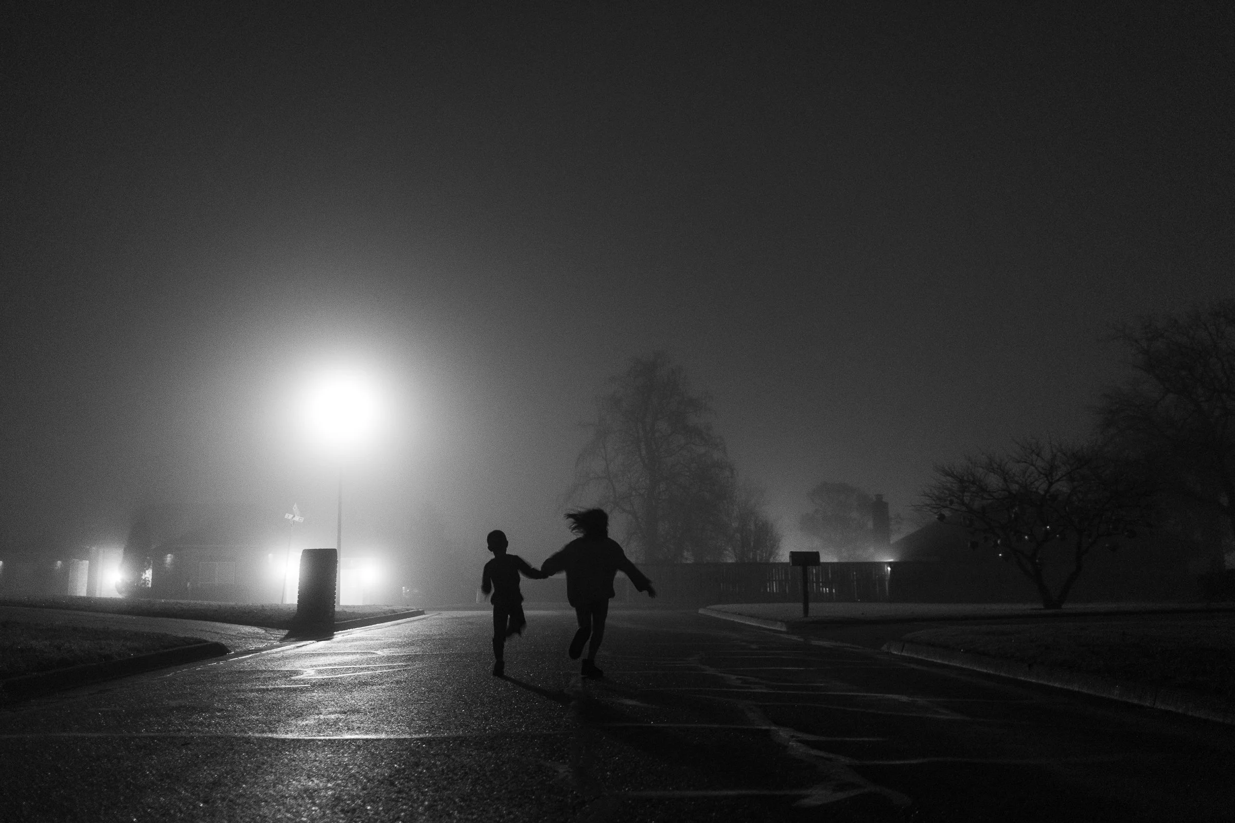 Two children running on a foggy, dark street at night illuminated by a bright streetlight, with trees and buildings in the background.