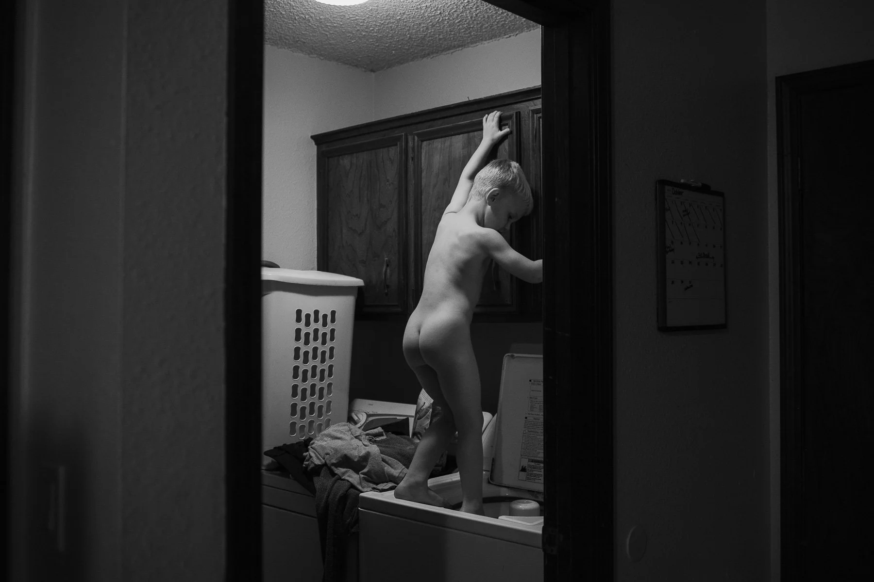 A young boy standing on a washing machine, reaching up to a cabinet in a laundry room, with laundry and a laundry basket visible.