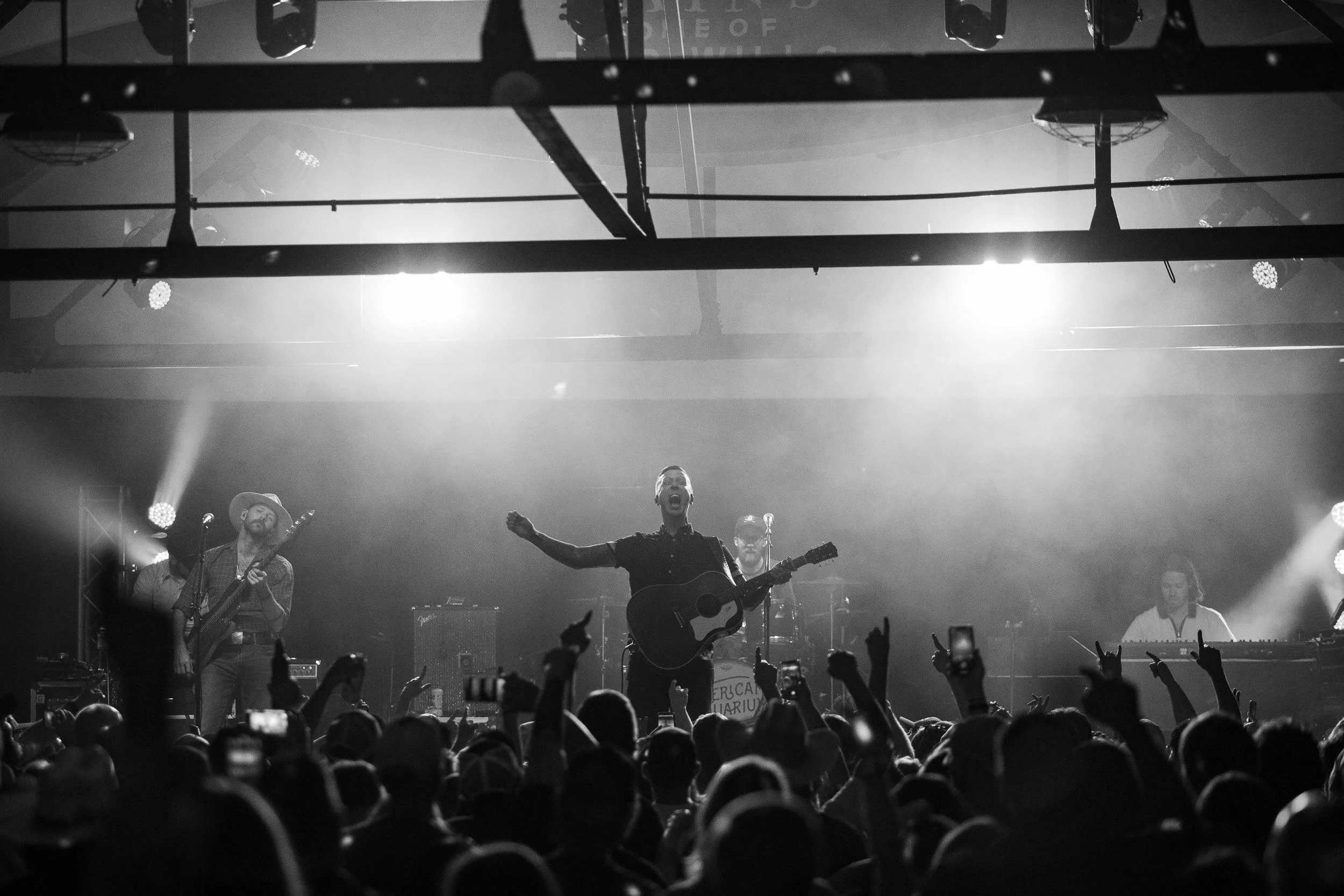 Black and white photo of a live musical performance with a band on stage and an audience in front watching.