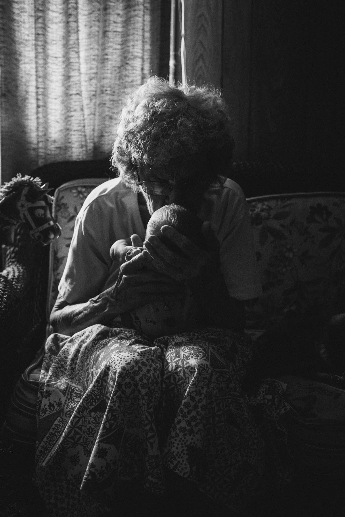 An elderly woman with curly hair and glasses sitting on a floral-patterned armchair, her head bowed and her hand resting on her cheek, in a dimly lit room with curtains and patterned fabric.