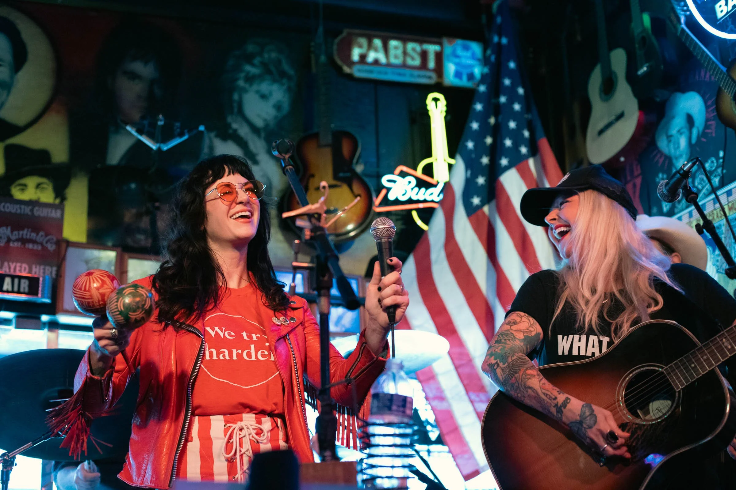 Two women performing on stage in a bar with American flags and neon signs, one with glasses and a red leather jacket holding maracas, the other with tattoos and a hat playing guitar, both smiling and laughing.