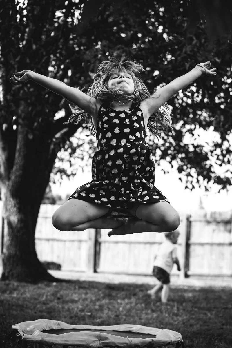 A young girl jumping in the air outdoors with arms outstretched and a big smile, wearing a dress with heart patterns. In the background, a boy runs near a fence under a large tree.