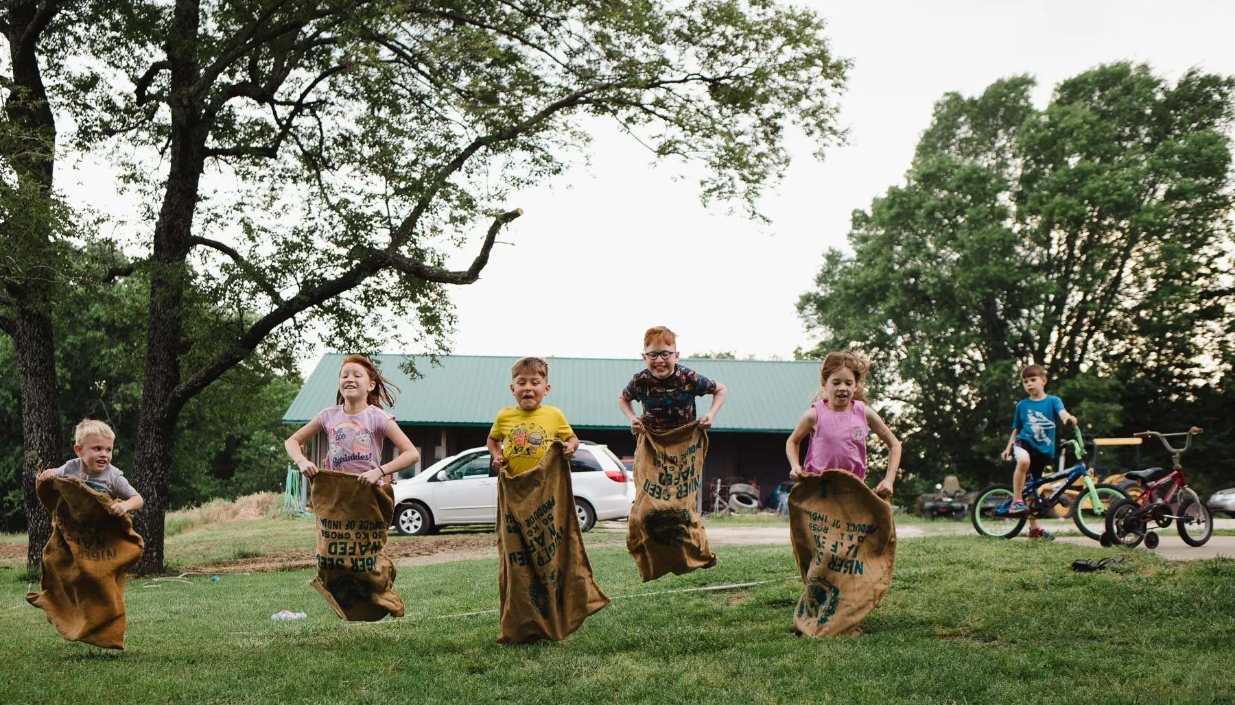 Children participating in a sack race outdoors during daytime, with two boys on bicycles in the background, trees, a building, and a grassy area.