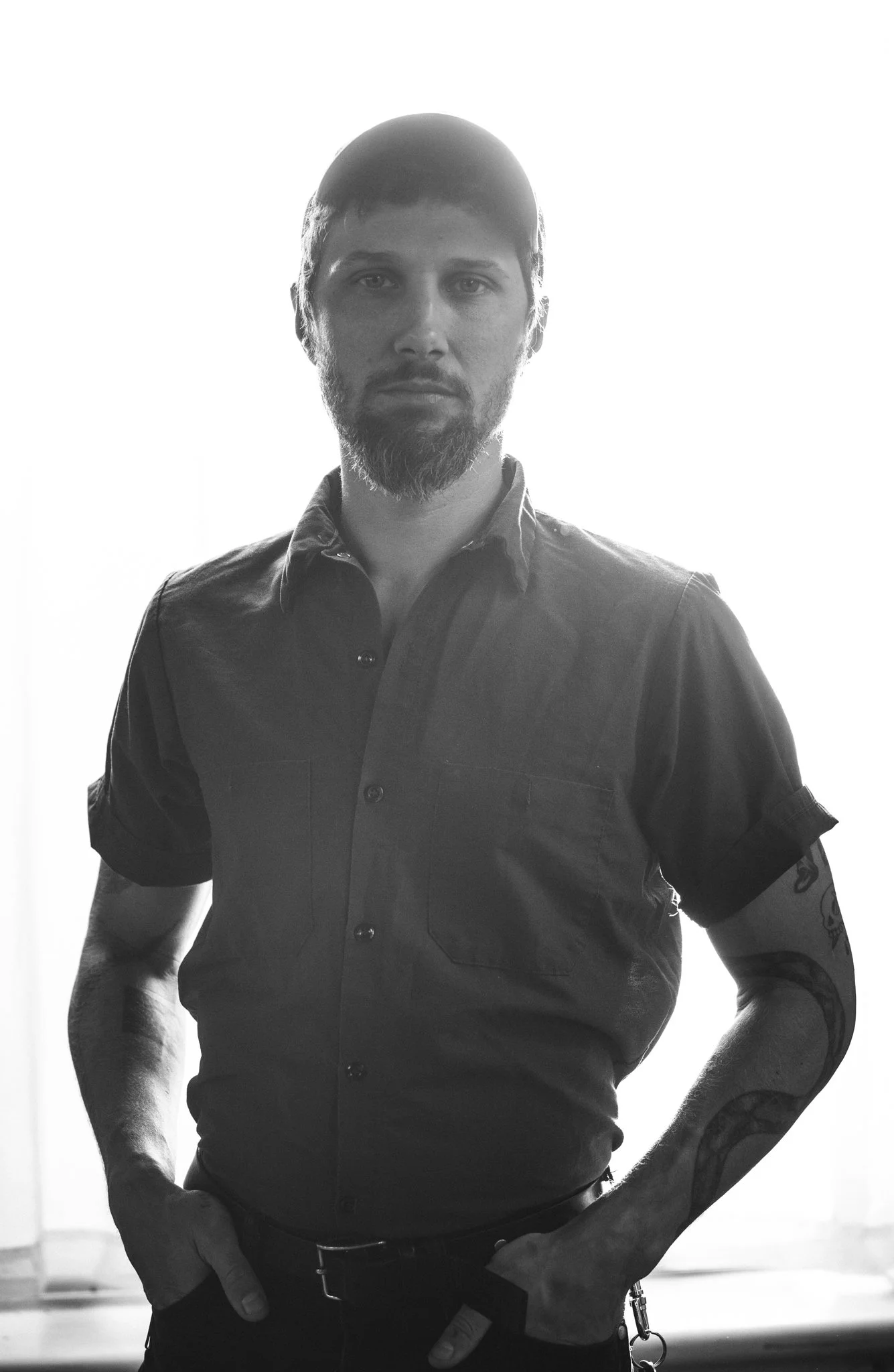 A man with a beard and tattoos, standing with hand in pocket, wearing a short-sleeved button-up shirt, backlit by bright light, creating a silhouette effect.