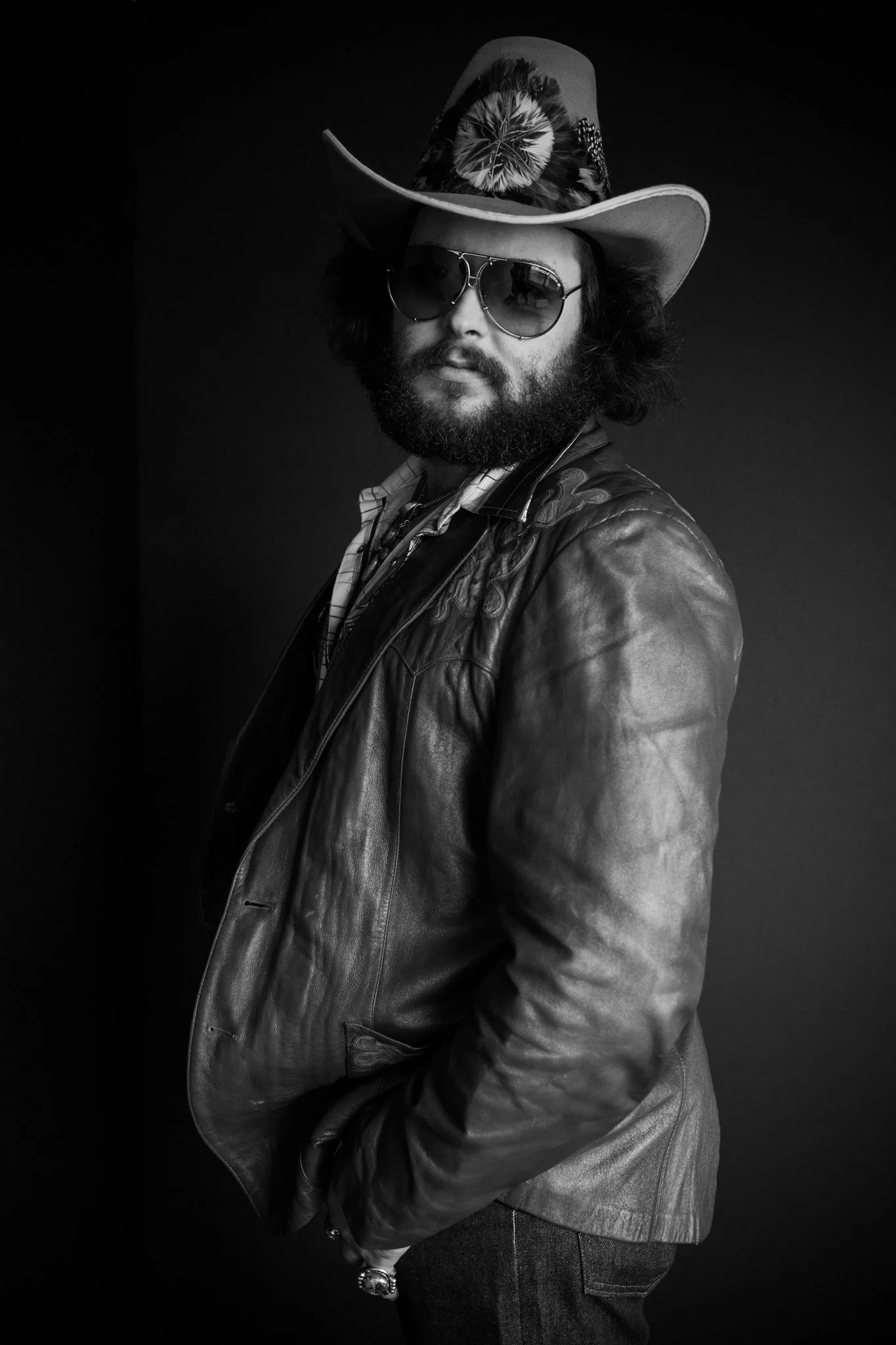 Black and white photo of a man wearing a cowboy hat with a floral pattern, sunglasses, a leather jacket, and a checkered shirt, against a dark background.