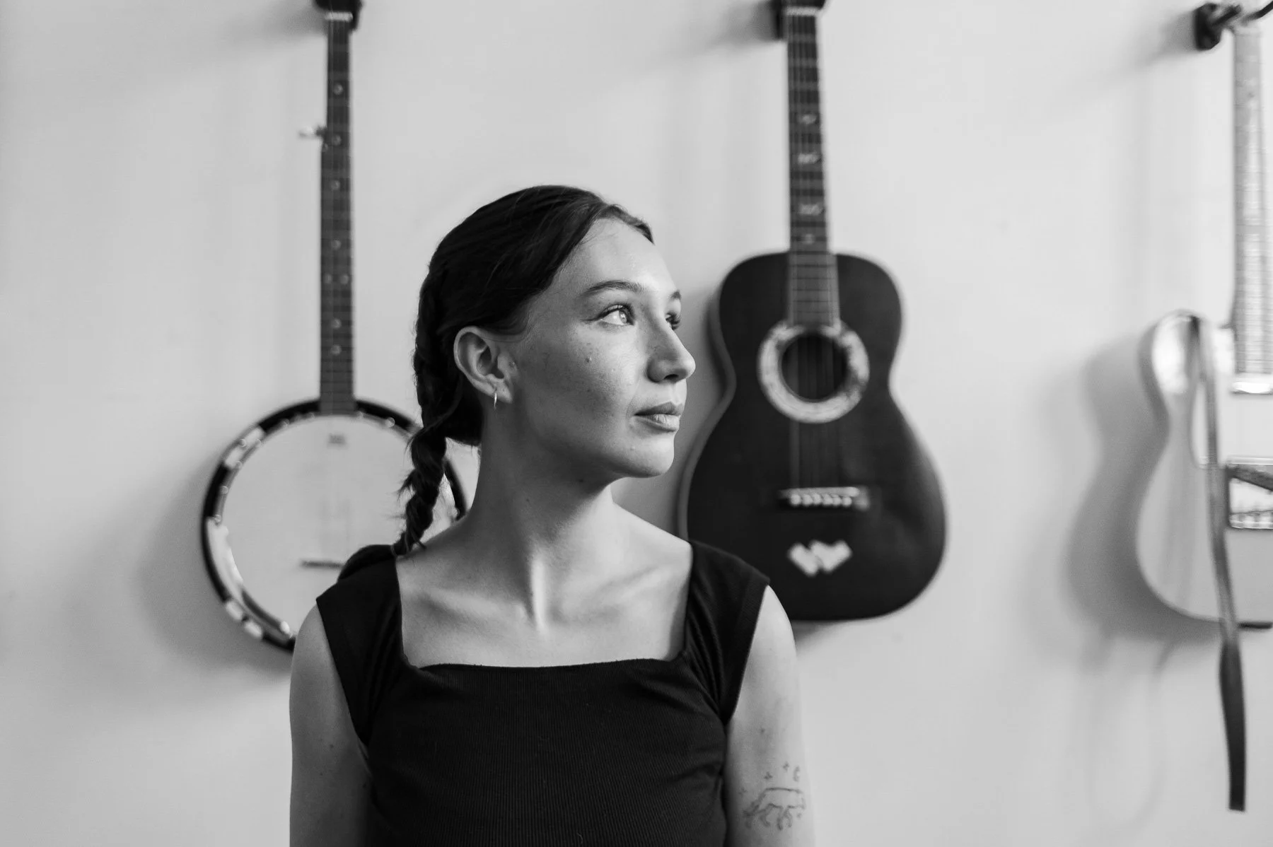 A woman with dark hair in a braid looks to the side in front of a wall with three guitars hanging, including an acoustic guitar and a banjo.