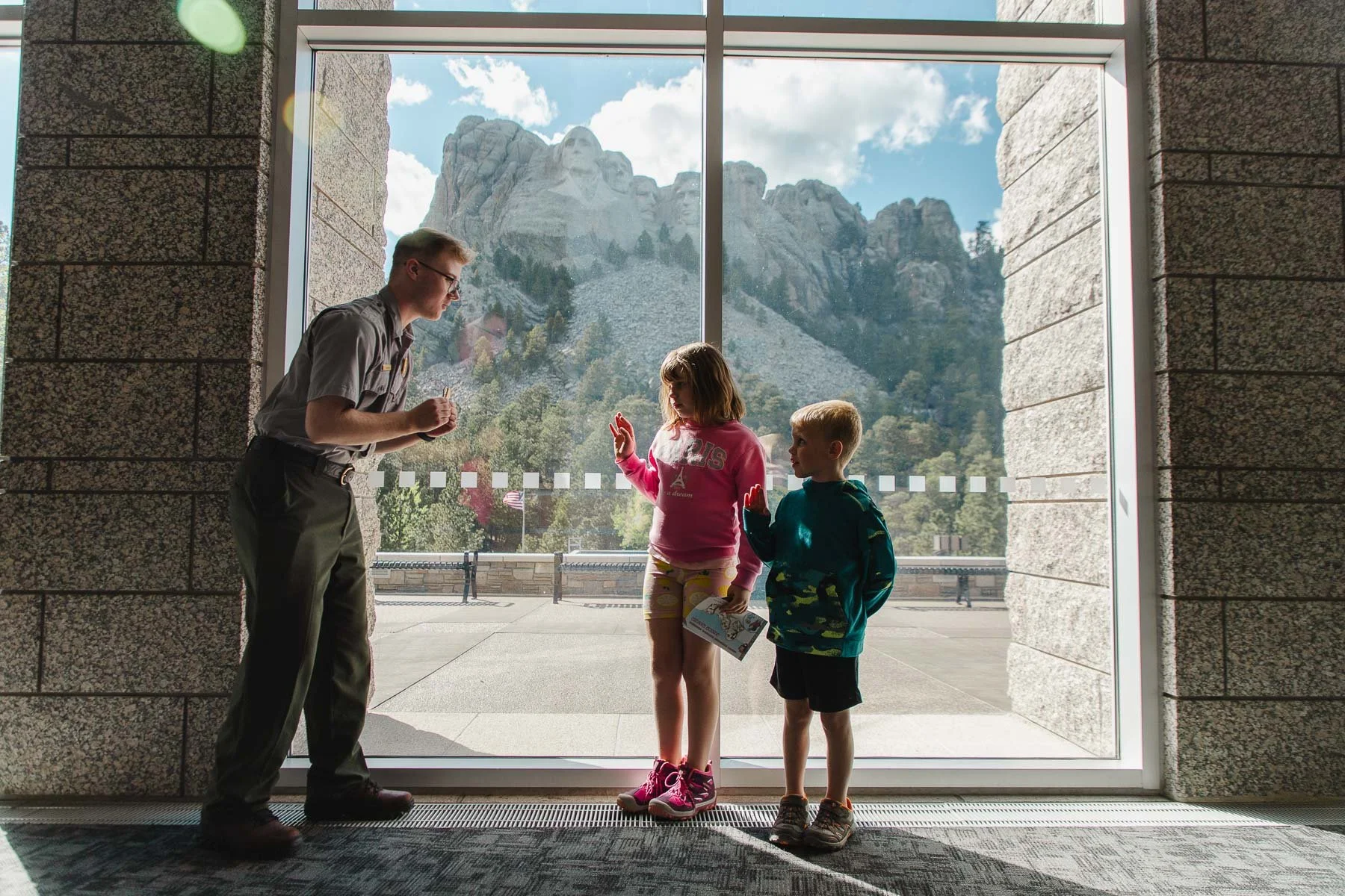 A park ranger shows children Mount Rushmore through a large glass window in a visitor center, with the sculpture of Mount Rushmore visible outside against a partly cloudy sky.