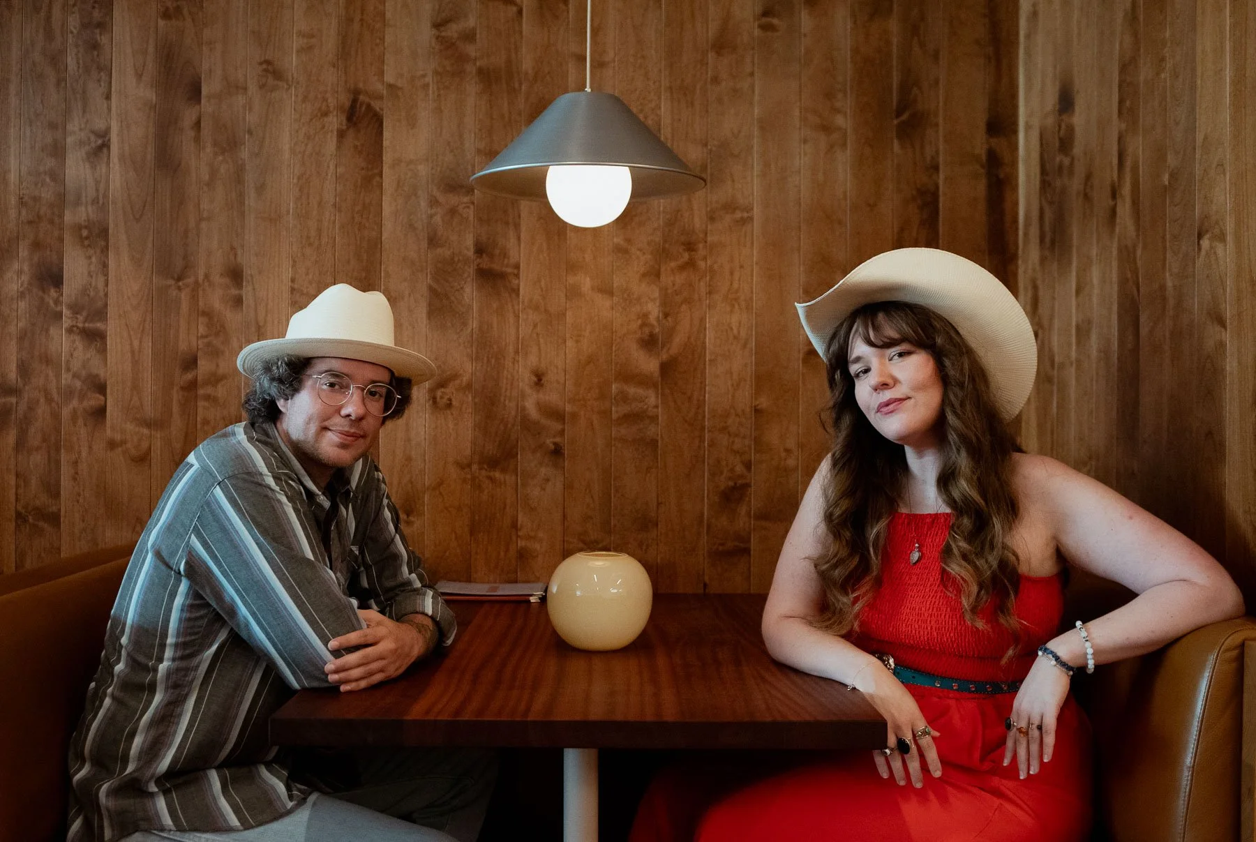 A man and a woman sitting at a wooden booth in a restaurant or cafe. The man is wearing a striped shirt, glasses, and a white hat, with arms crossed on the table. The woman is wearing a red dress, white hat, and jewelry, with her arm on the table and