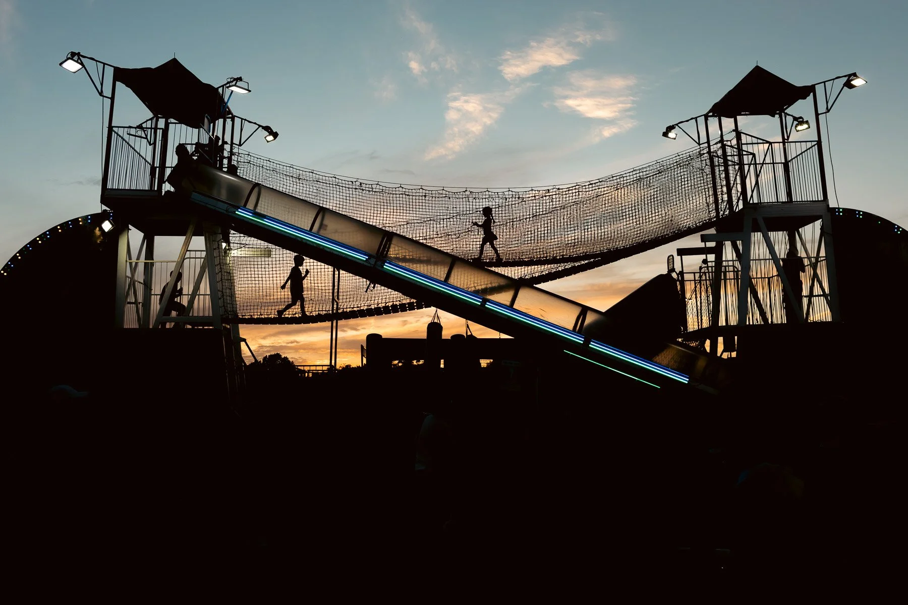 Silhouette of children playing on a carnival amusement park ride at sunset, including a slide and a climbing net structure with towers.