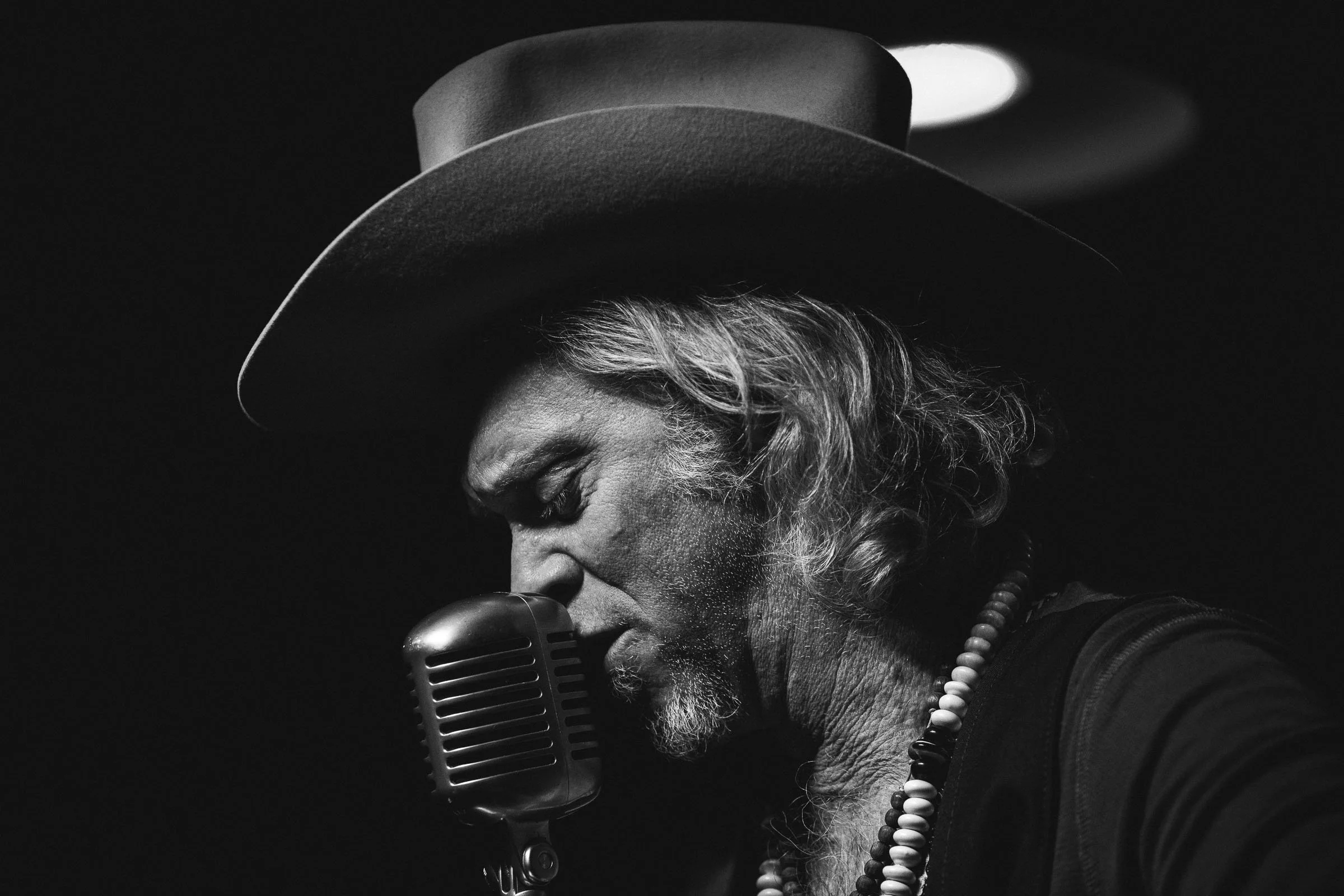 A black and white photo of a man with long hair and a beard, wearing a wide-brimmed hat and beads, singing into a vintage microphone with his eyes closed.