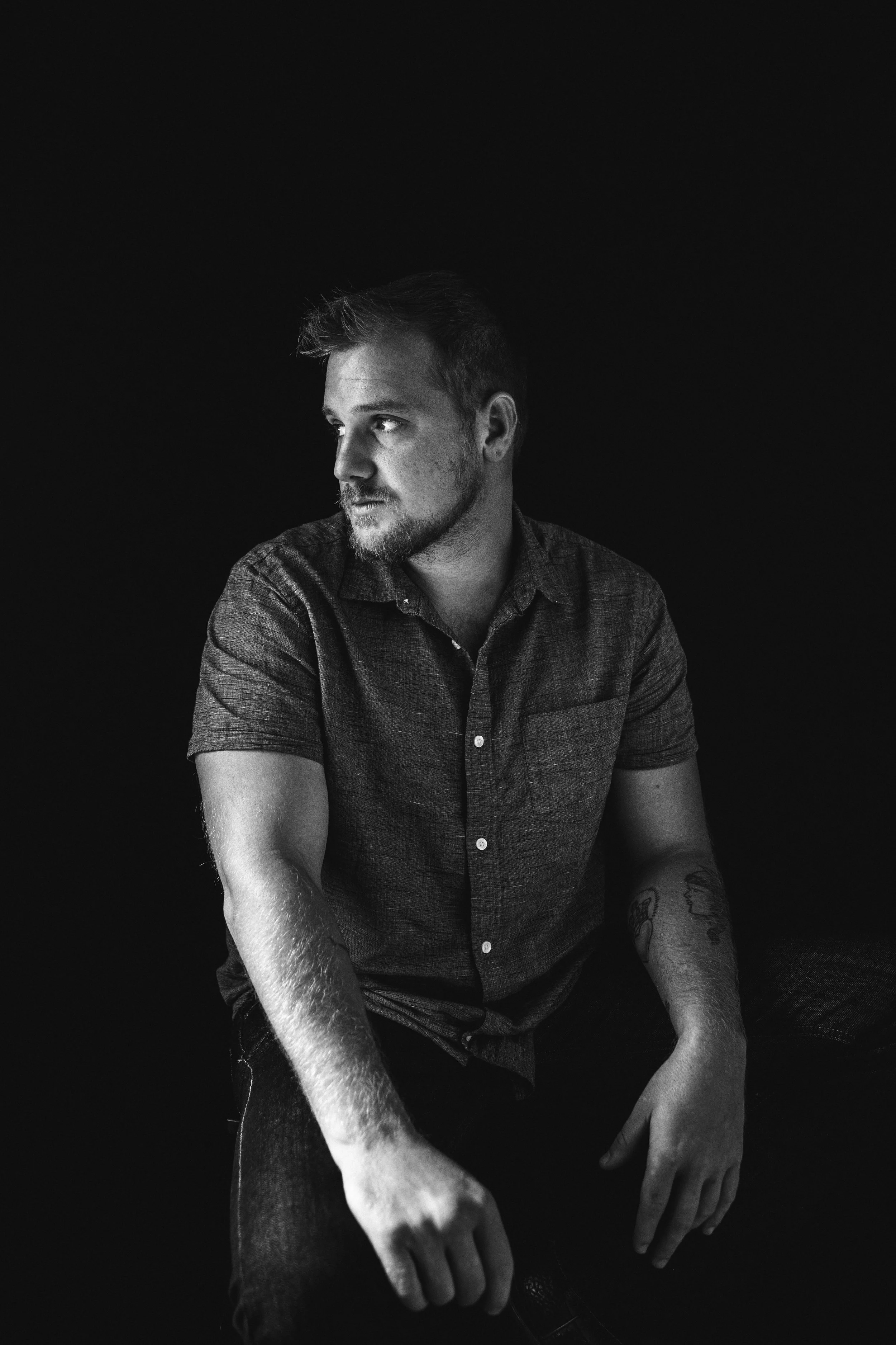 Black and white portrait of a man with a beard, wearing a short-sleeve button-up shirt, sitting against a black background, looking to his left.