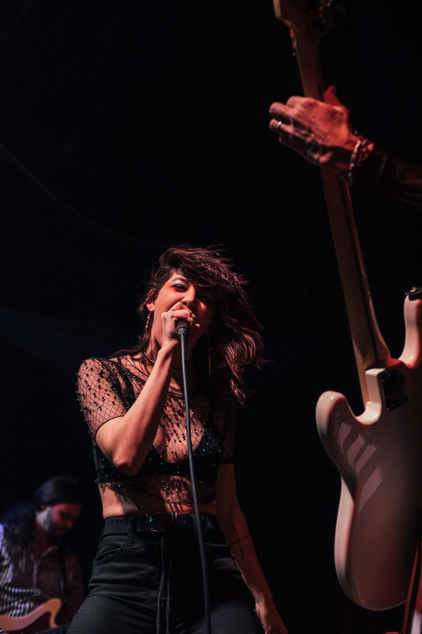 A woman singing into a microphone on stage with a guitarist in the background.