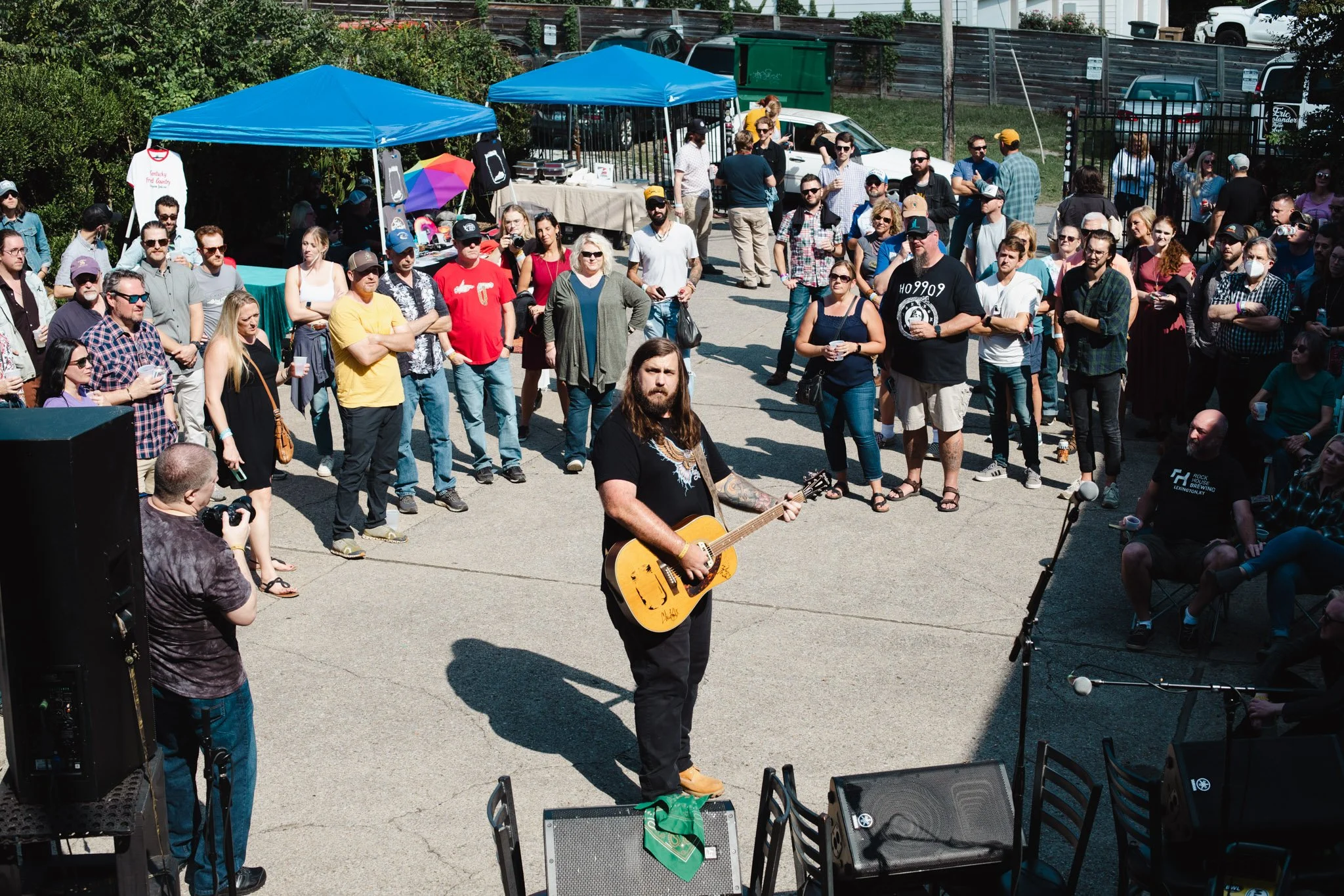 A man with long hair and a beard playing an acoustic guitar in front of a crowd at an outdoor event. The audience is standing and sitting, some wearing sunglasses, with tents and vendor booths in the background.