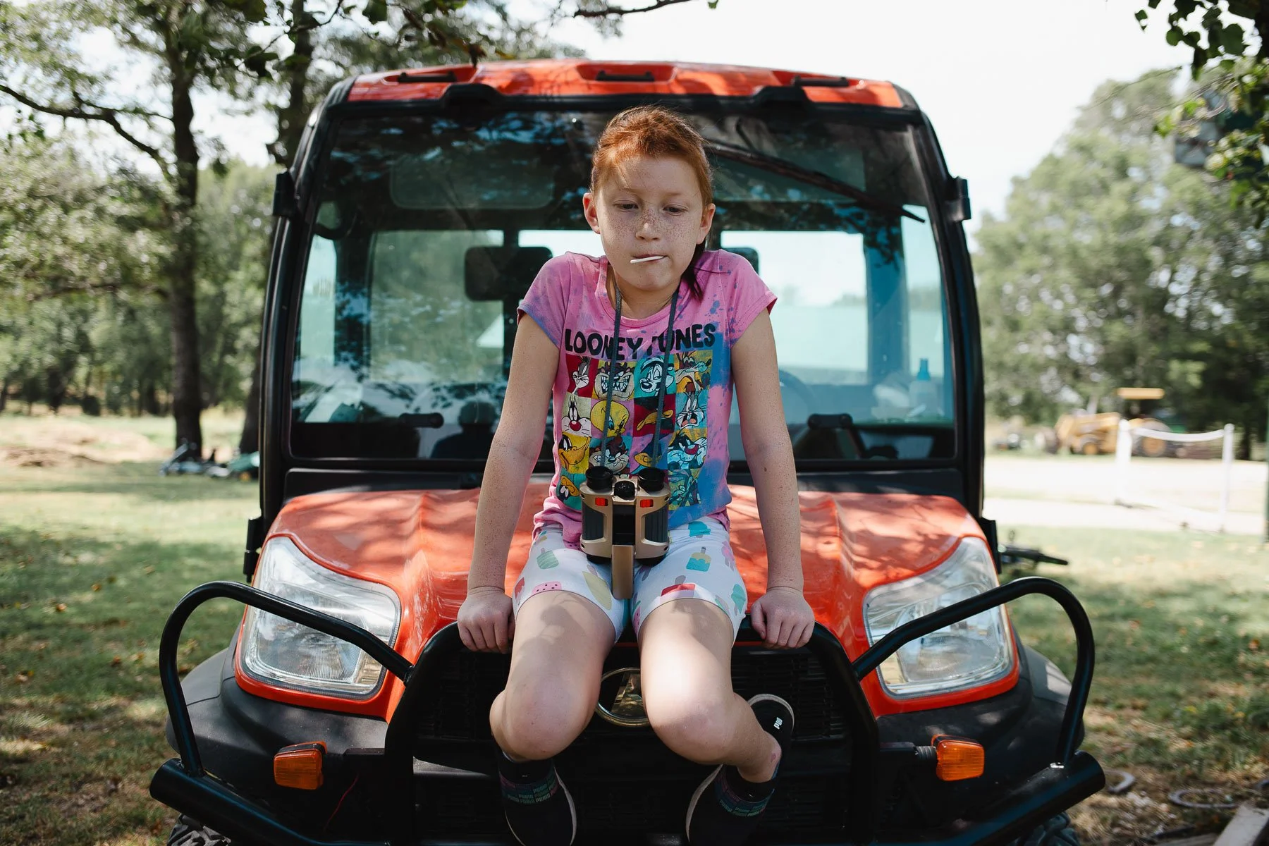 A young girl sitting on the front of a small orange utility vehicle, holding binoculars around her neck, outdoors in a park with trees and a grassy field.