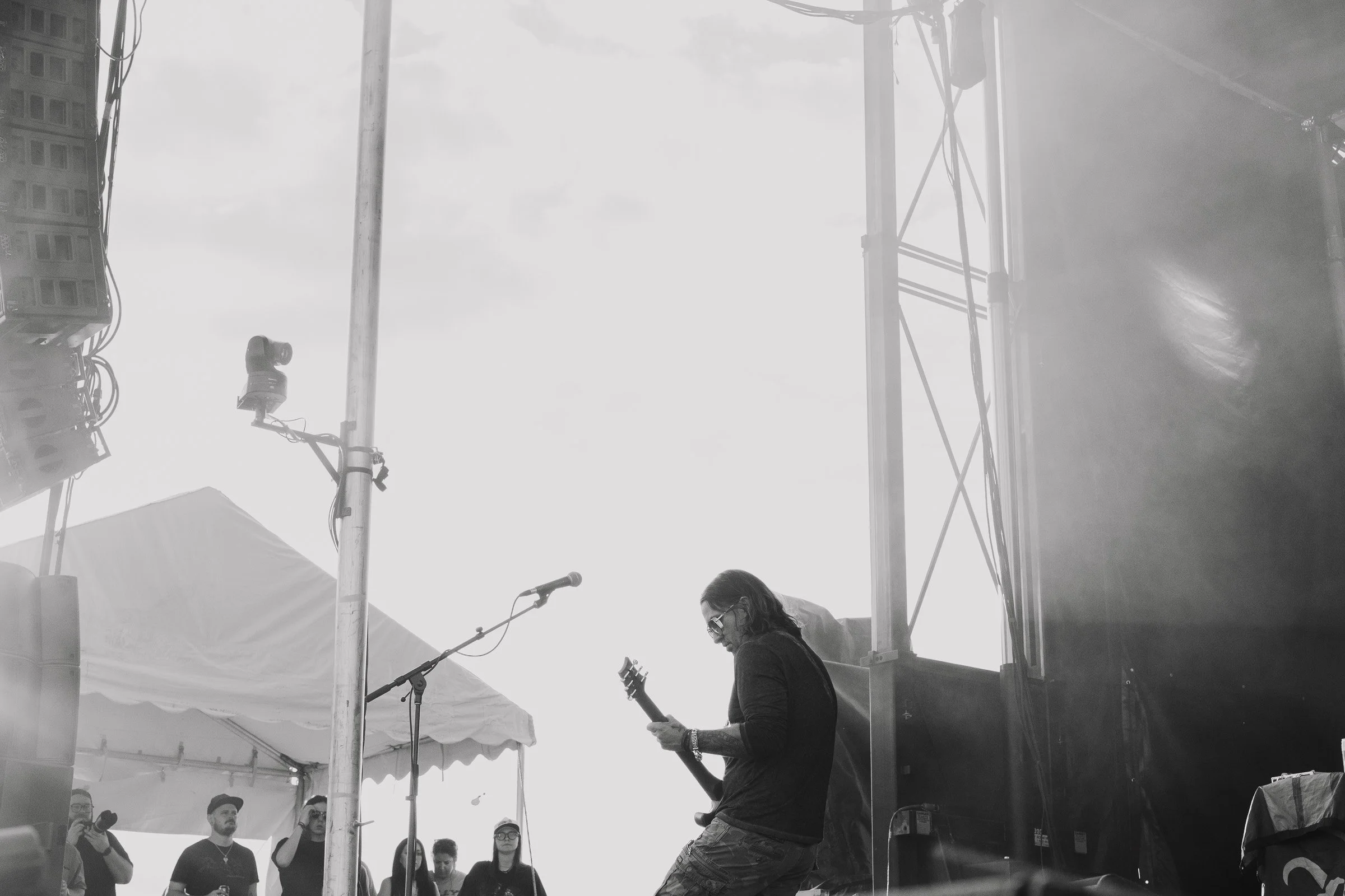 Musician playing guitar on stage at an outdoor concert, with several audience members watching under a tent, black and white image.
