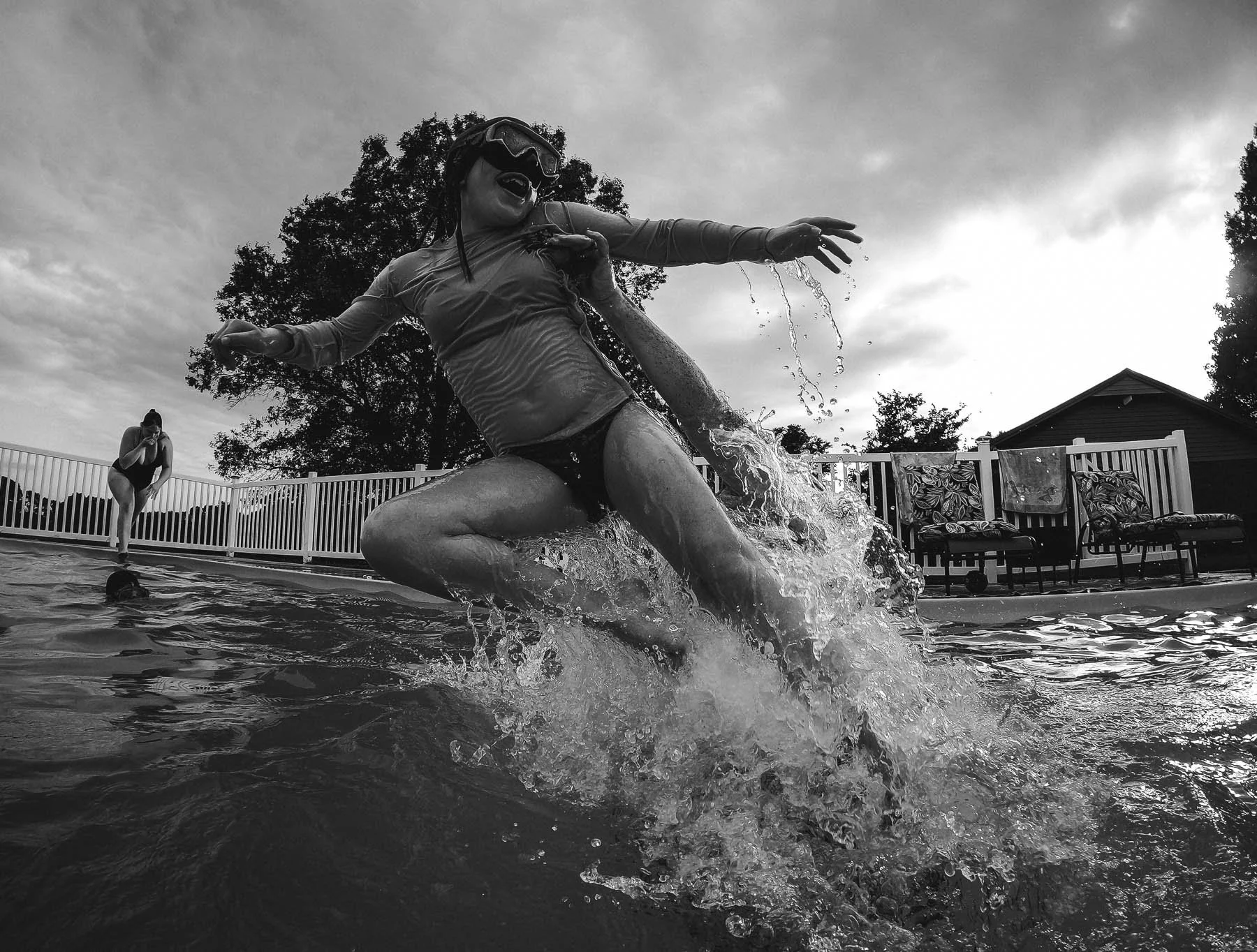Person in swim goggles and swimwear jumping into a pool, splashing water, with another person in a swimsuit standing at the poolside, and outdoor furniture and trees in the background.