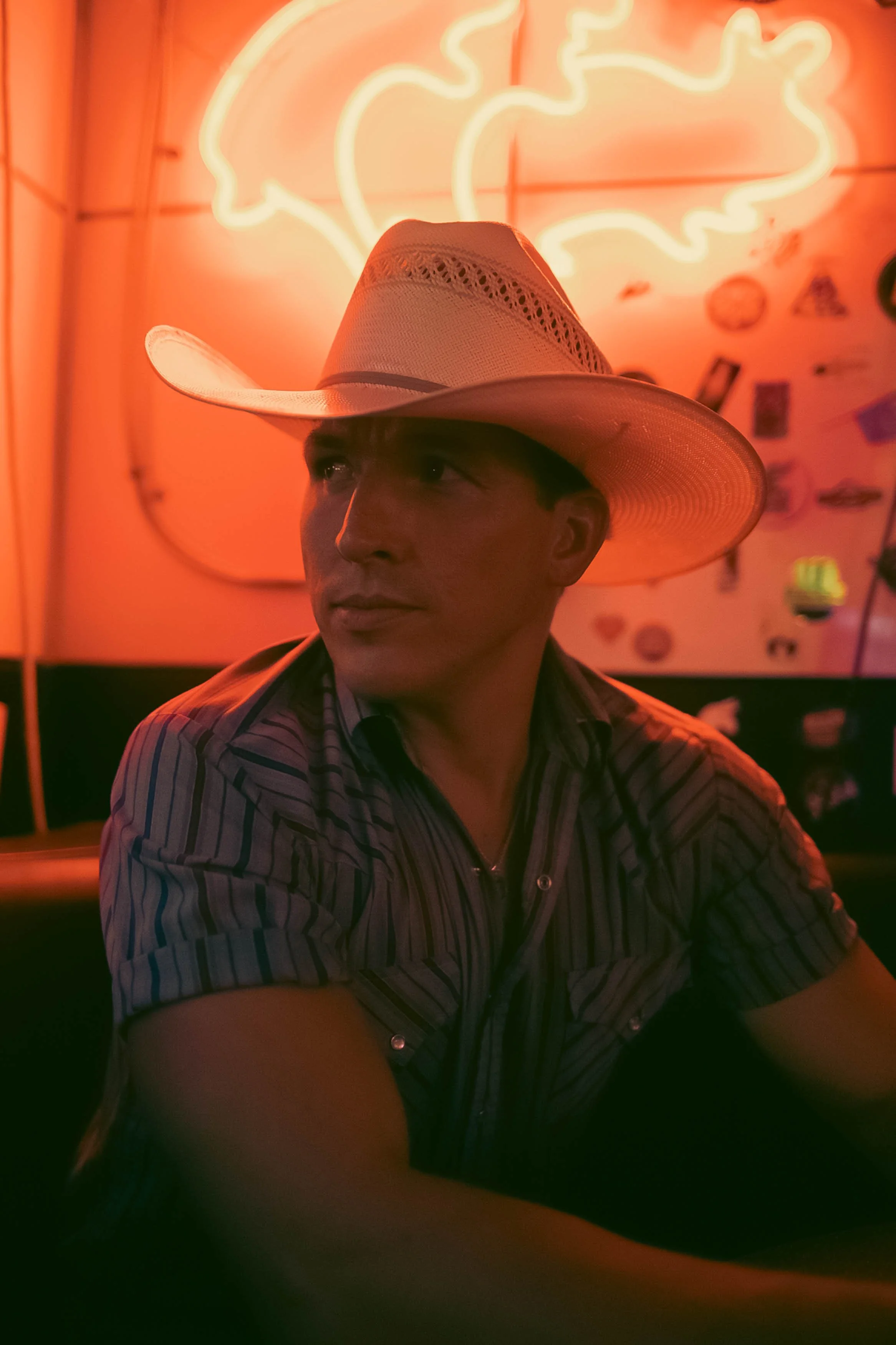 A man wearing a cowboy hat sitting in a dimly lit room with orange neon lights behind him.